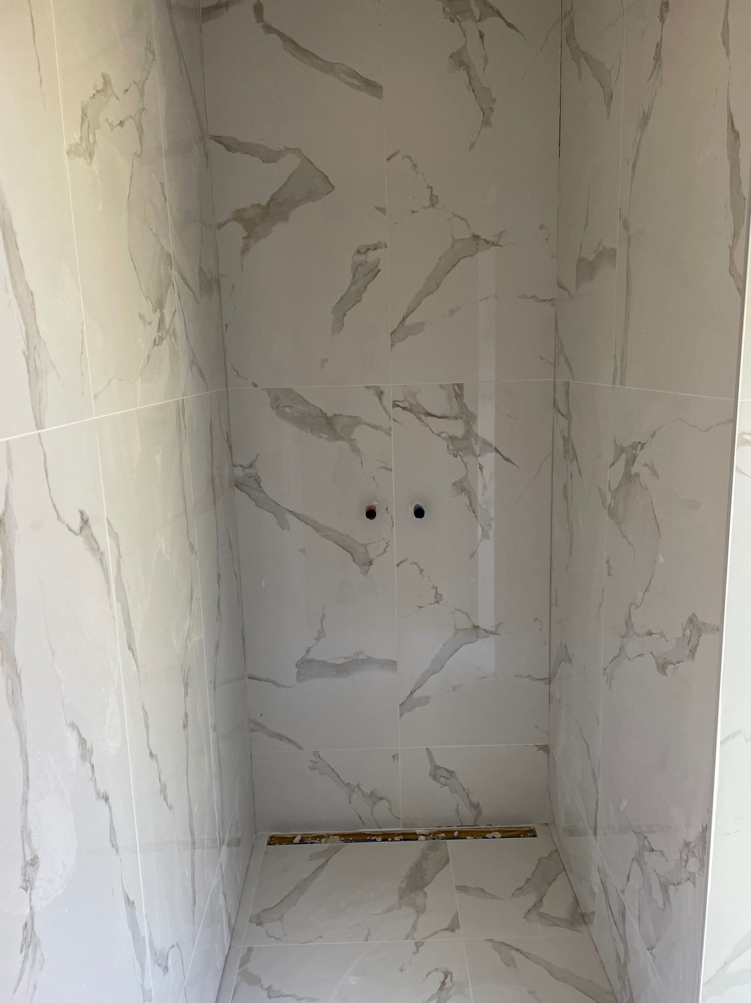 Wnętrze kabiny prysznicowej wyłożonej białymi płytkami imitującymi marmur, widoczne otwory na baterię i odpływ liniowy w podłodze, prace w trakcie.