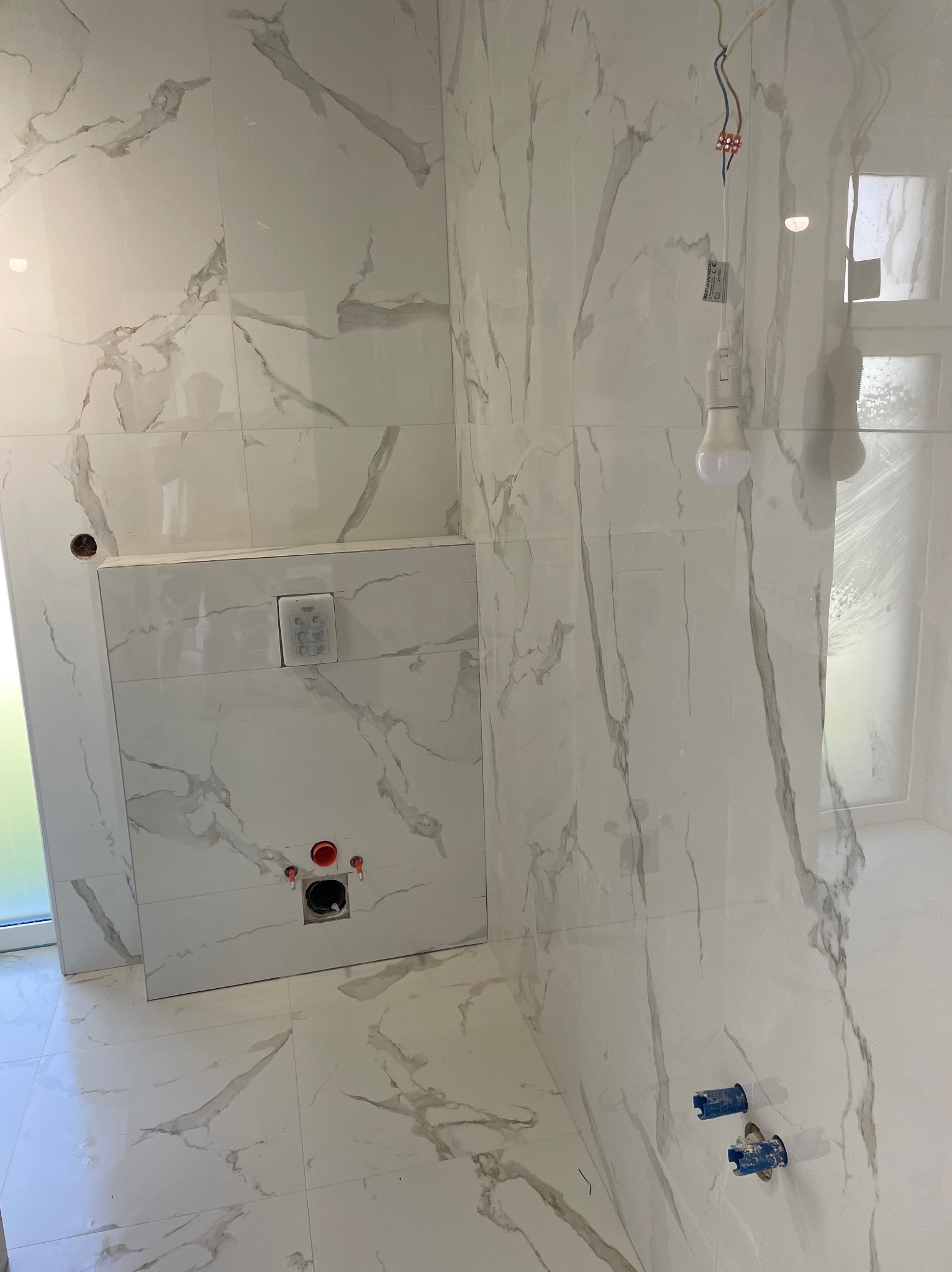 Wnętrze łazienki w trakcie wykańczania, widoczne białe płytki imitujące marmur na ścianach i podłodze, podtynkowa spłuczka WC z przyciskiem, wystające przewody instalacji wodnej i elektrycznej...