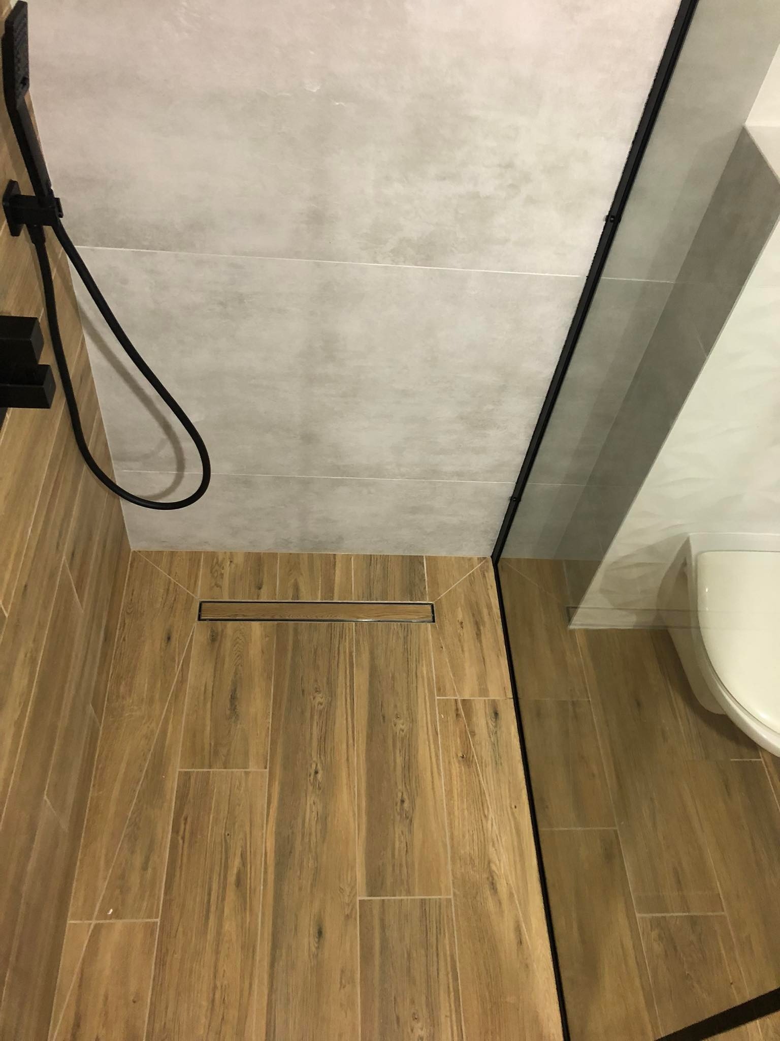 Nowoczesna kabina prysznicowa walk-in z odpływem liniowym, wykończona płytkami imitującymi drewno, czarne elementy armatury.