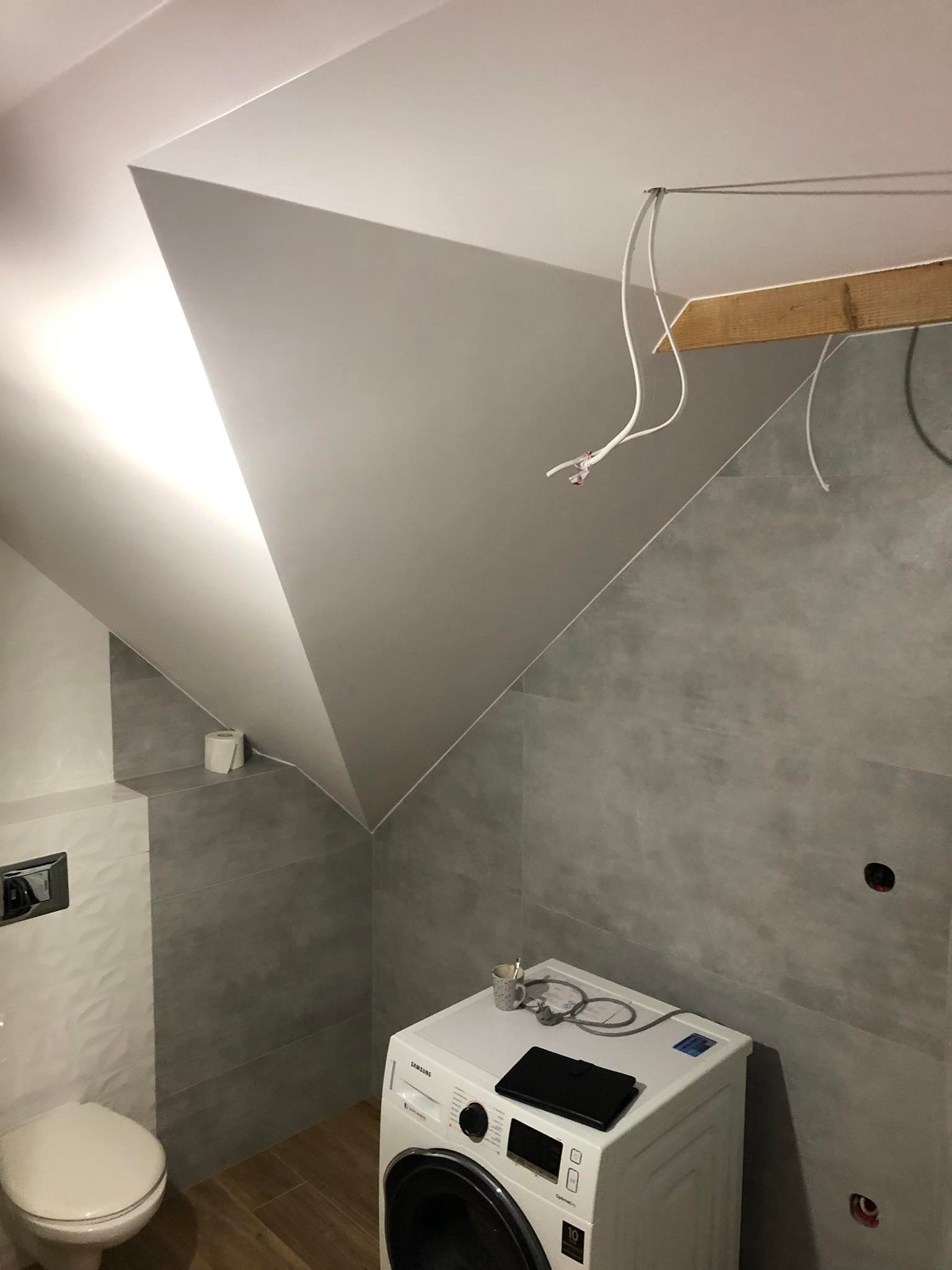 Łazienka na poddaszu w trakcie remontu: szare płytki, biała pralka z czarnym tabletem na górze, instalacja elektryczna zwisająca z sufitu, drewniana belka podtrzymująca przewody.
