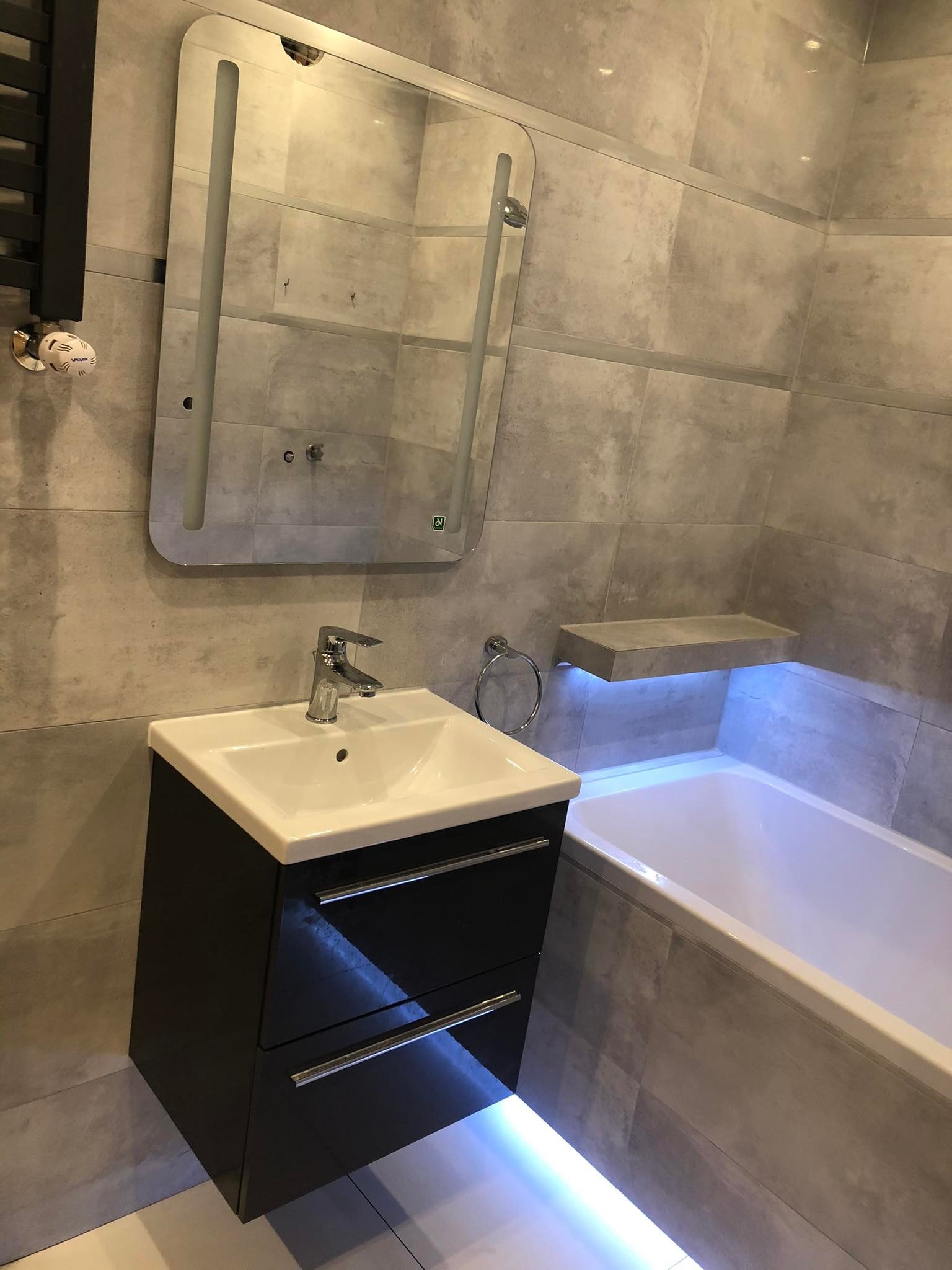 Nowoczesna łazienka z szarymi płytkami, podświetlaną wanną i szafką pod umywalką z lustrem.