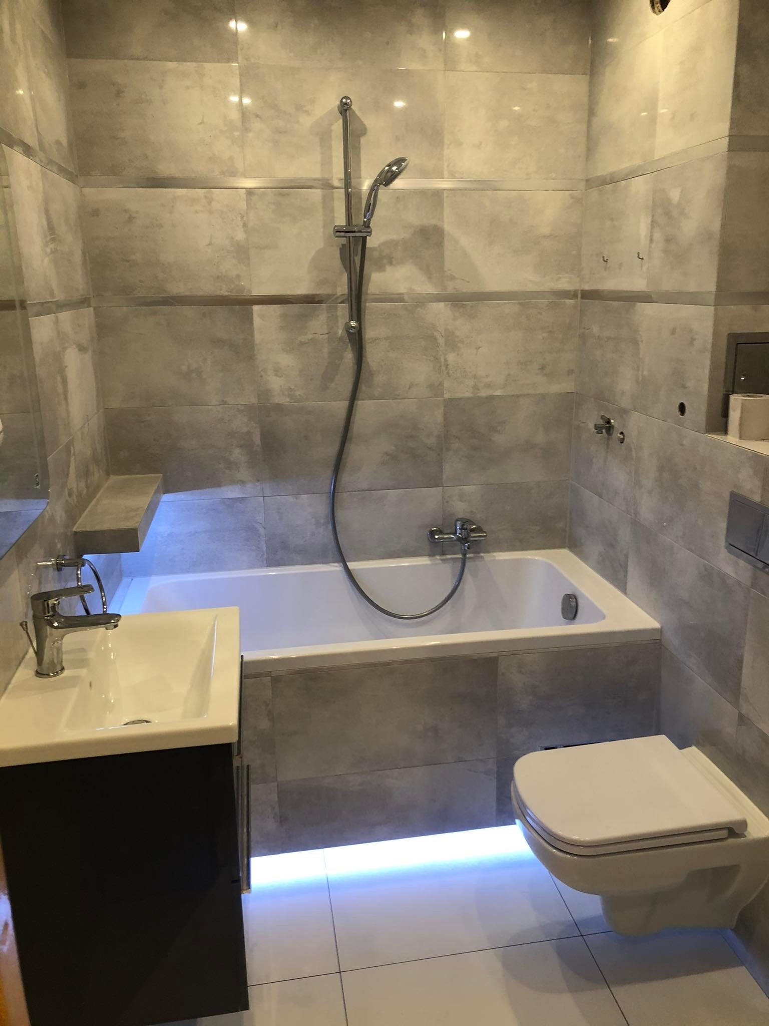 Ukończona łazienka z szarymi, kwadratowymi płytkami, podświetlaną wanną i umywalką z szafką oraz wiszącą toaletą.