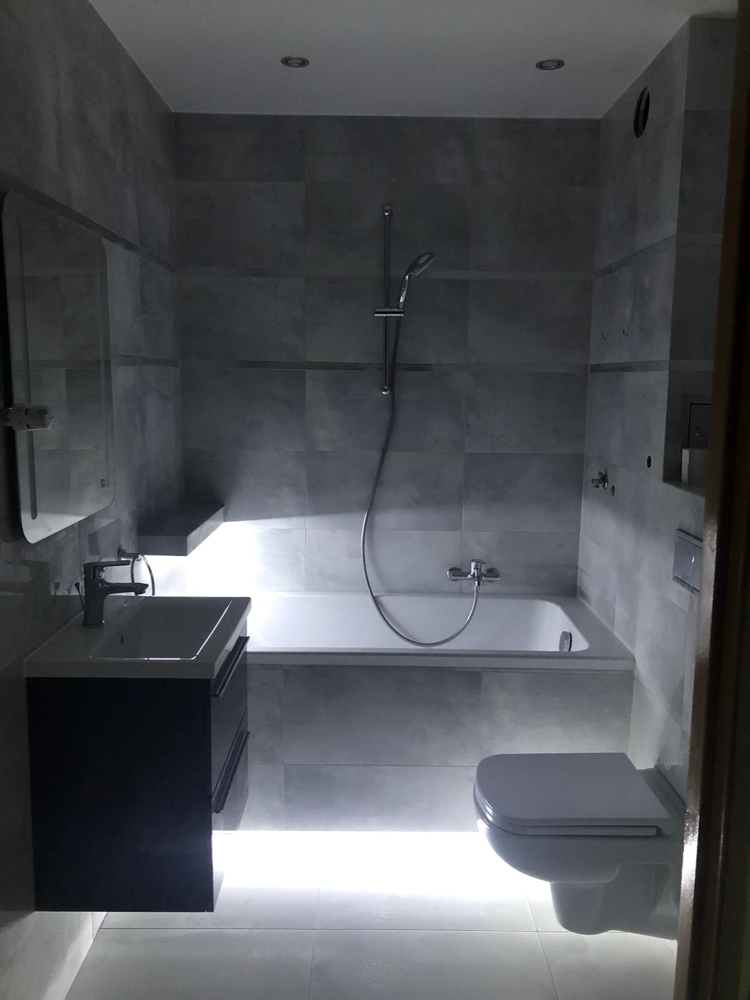 Nowoczesna łazienka w odcieniach szarości z podświetleniem LED pod umywalką i toaletą, płytki imitujące beton, minimalistyczna armatura.