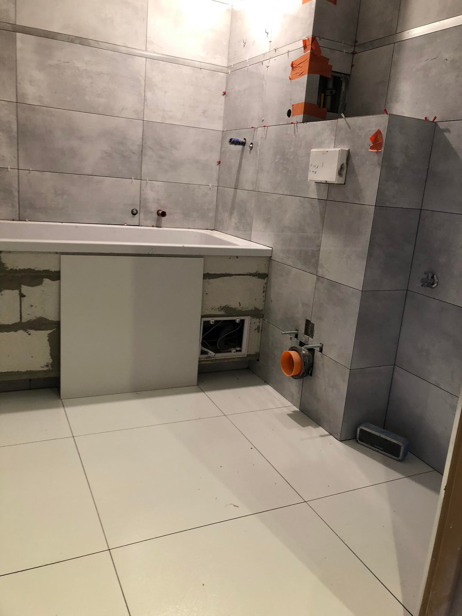 Łazienka w trakcie remontu: szare płytki na ścianach z pomarańczowymi zabezpieczeniami, biała wanna z obudową, białe płytki na podłodze, widoczne rury i elementy instalacji.