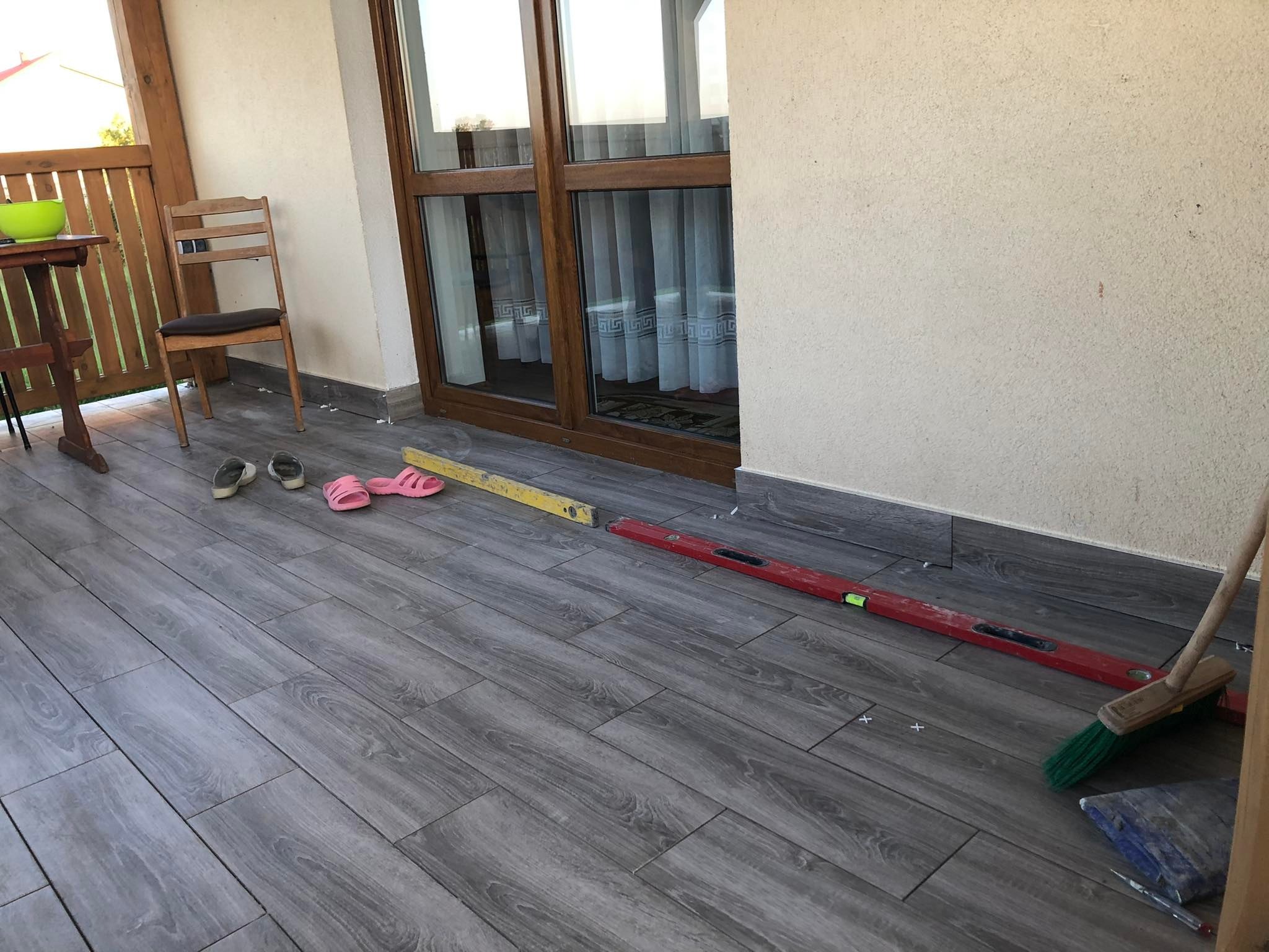 Układanie szarych płytek imitujących drewno na balkonie, widoczne poziomice, krzyżyki dystansowe, szczotka do zamiatania i narzędzia glazurnicze.