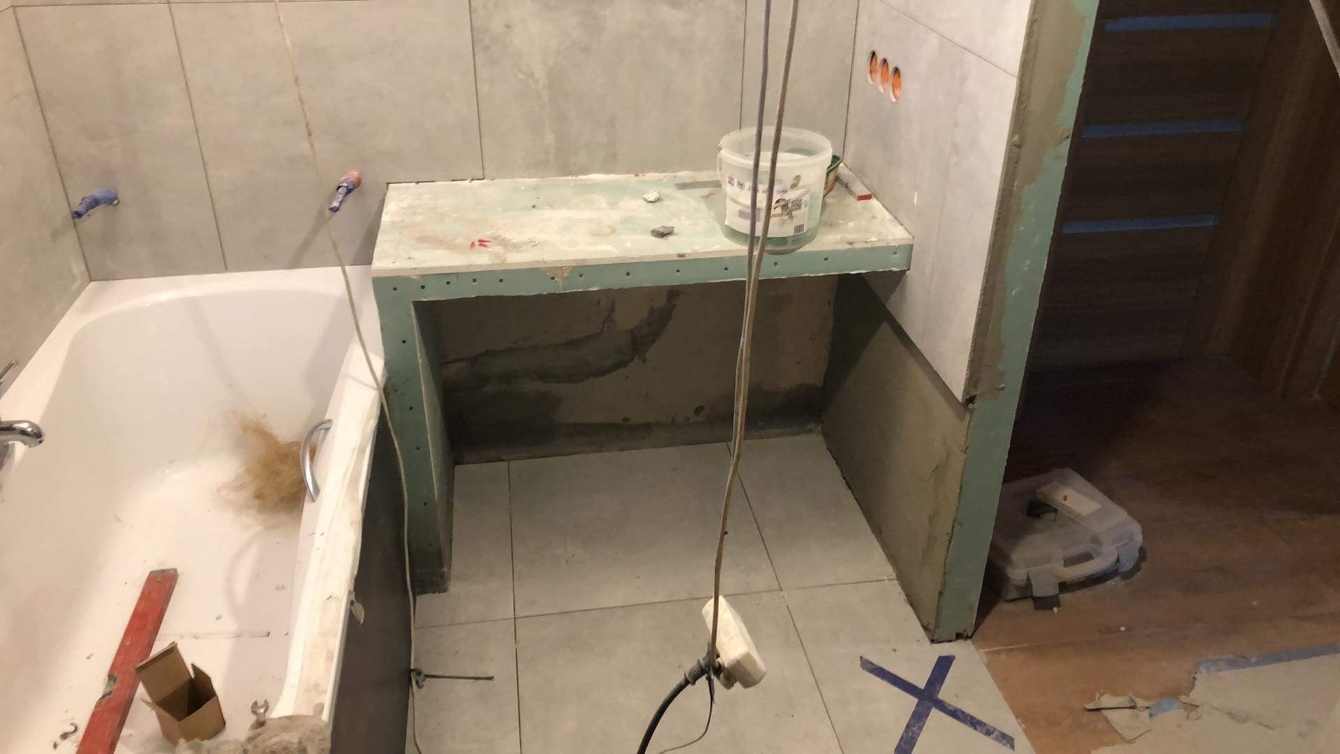 Łazienka w trakcie remontu: obudowa pod umywalkę z płyt kartonowo-gipsowych, wanna z pozostałościami po remoncie, płytki na ścianach, widoczne ślady prac wykończeniowych.