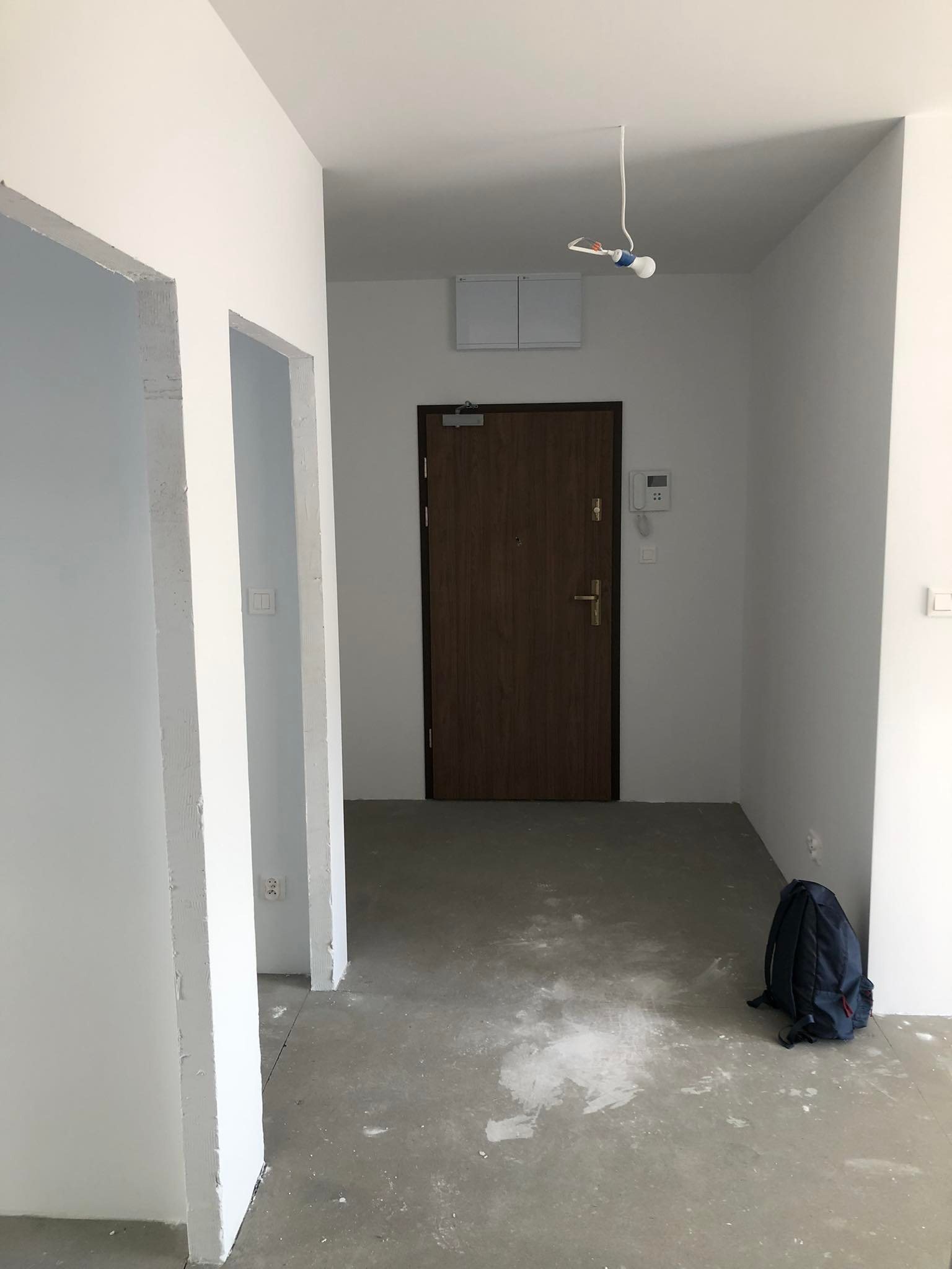 Surowe wnętrze mieszkania w trakcie remontu: widoczne białe, świeżo pomalowane ściany, surowy beton na podłodze, drewniane drzwi wejściowe i niebieski plecak leżący na podłodze.