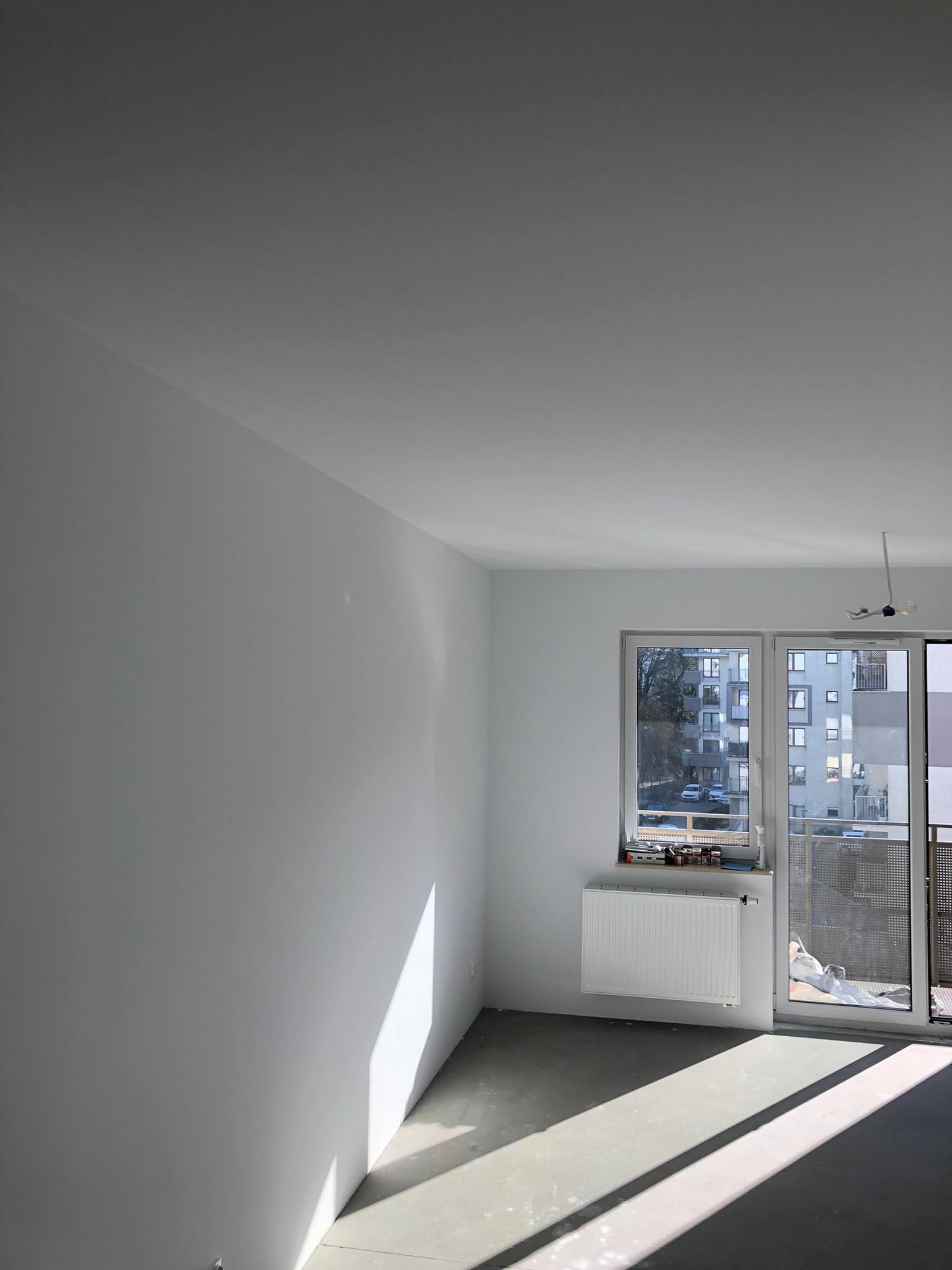 Wnętrze pokoju po świeżym malowaniu na biało, widoczne promienie słoneczne wpadające przez okno balkonowe, grzejnik pod oknem, surowa podłoga.