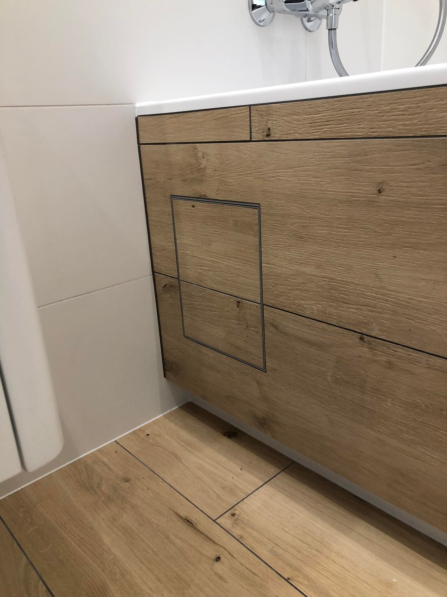 Nowoczesna zabudowa wanny z drewnopodobną okładziną, z ukrytymi drzwiczkami rewizyjnymi, dopasowana do podłogi z paneli imitujących drewno, białe płytki na ścianach.