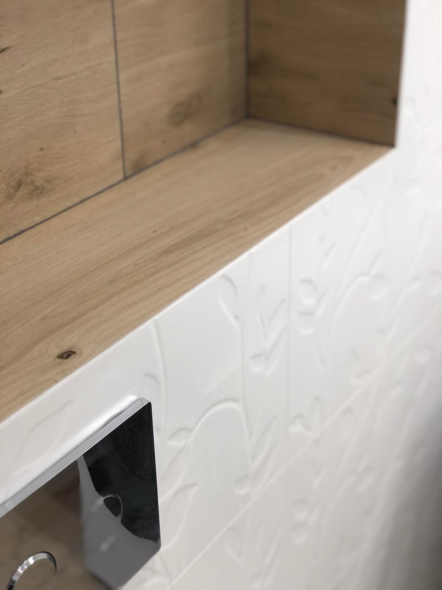 Narożnik w łazience z drewnianą półką i białą ścianą z płytek z delikatnym wzorem roślinnym, widoczny fragment chromowanego przycisku spłuczki.