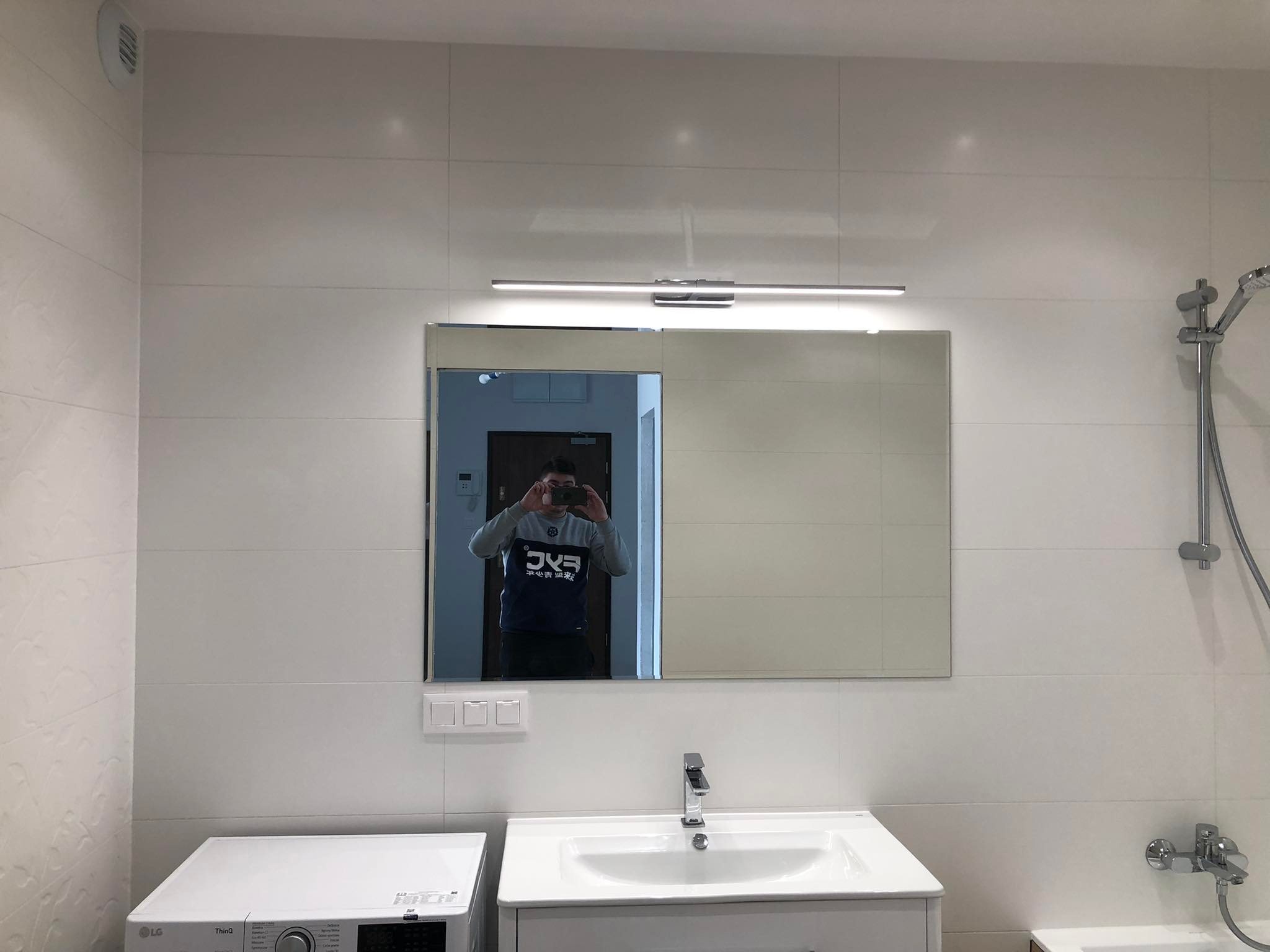 Biała łazienka z umywalką, lustrem podświetlanym listwą LED i pralką, widoczne białe płytki na ścianach, bateria umywalkowa chromowana, odbicie osoby robiącej zdjęcie w lustrze.