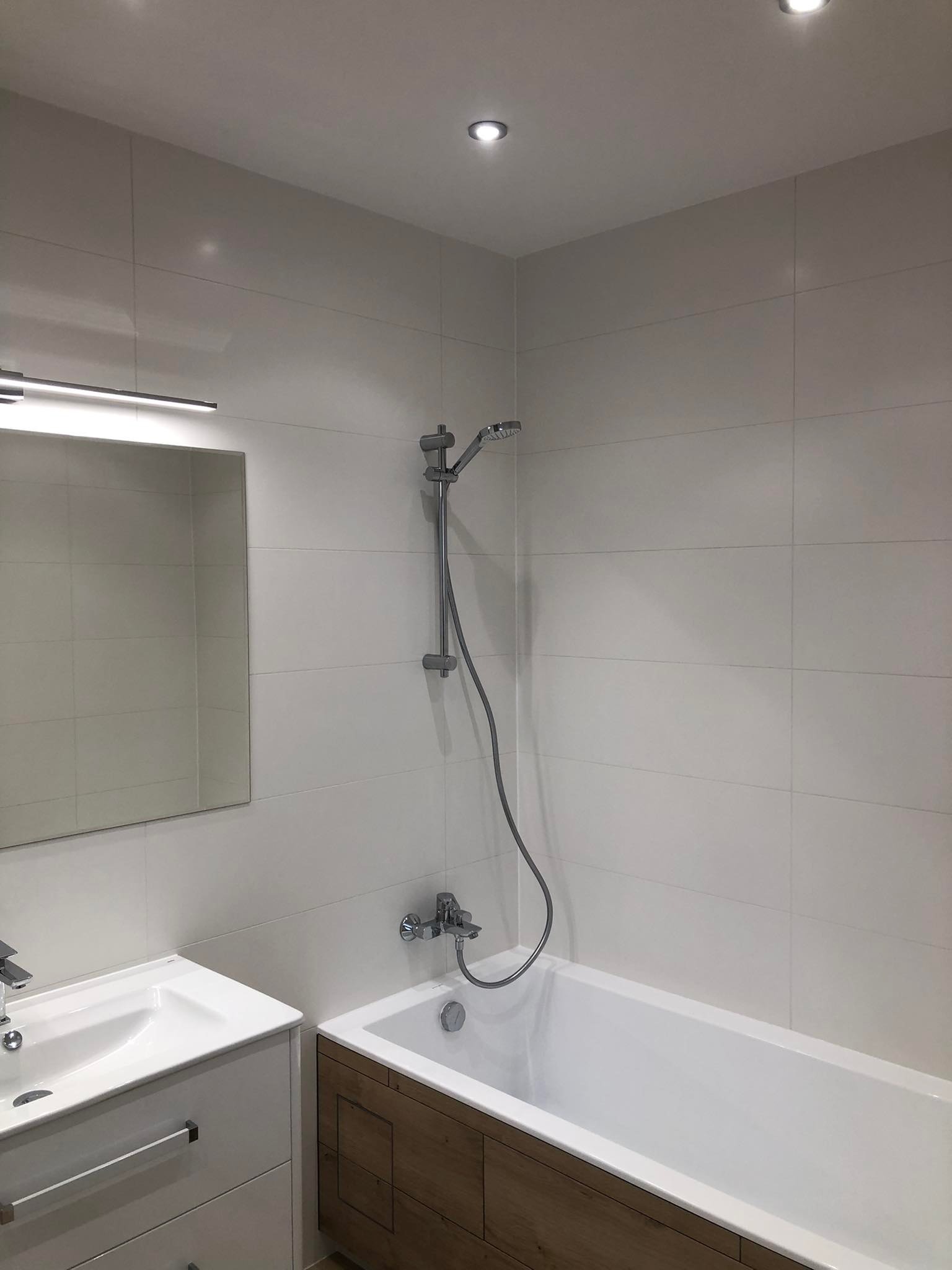 Nowoczesna łazienka z białymi płytkami, drewnianą zabudową wanny i minimalistyczną armaturą, oświetlona punktowo.