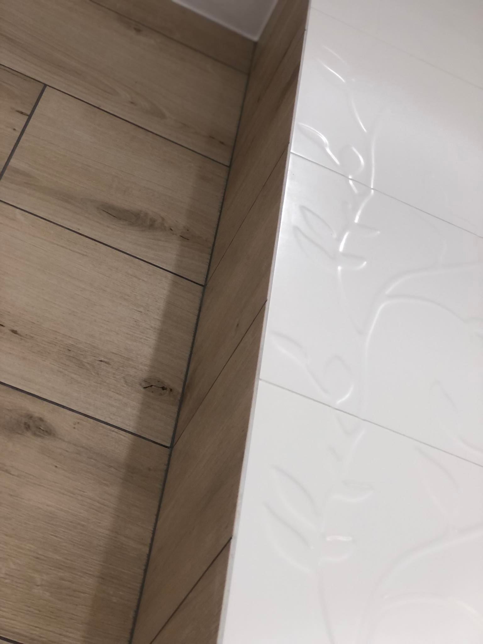 Połączenie drewnopodobnych płytek podłogowych i białych, zdobionych płytek ściennych w łazience, widoczne fugi.