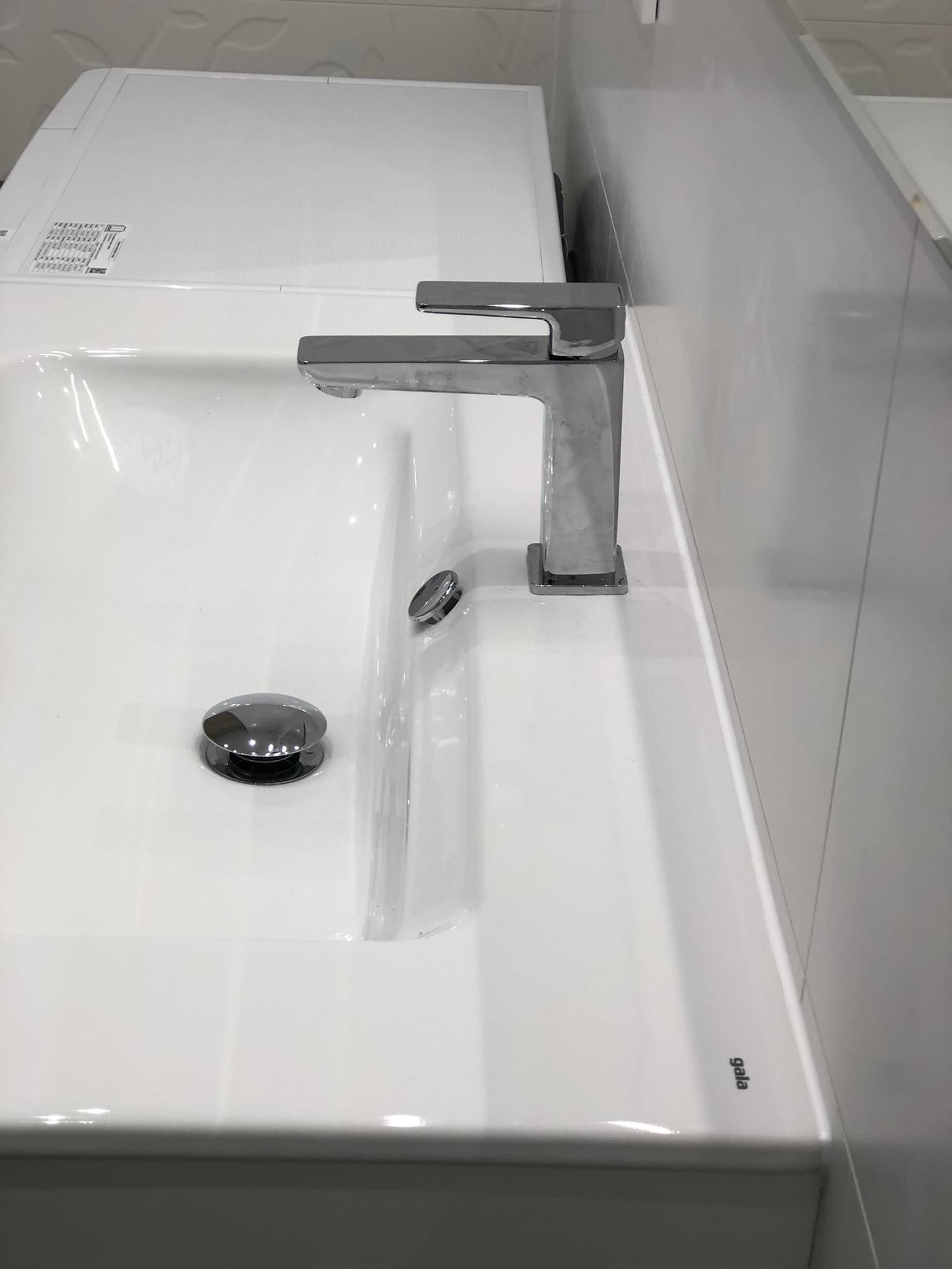 Biała umywalka z chromowaną baterią i odpływem typu click-clack, widoczne białe płytki na ścianie i pralka.