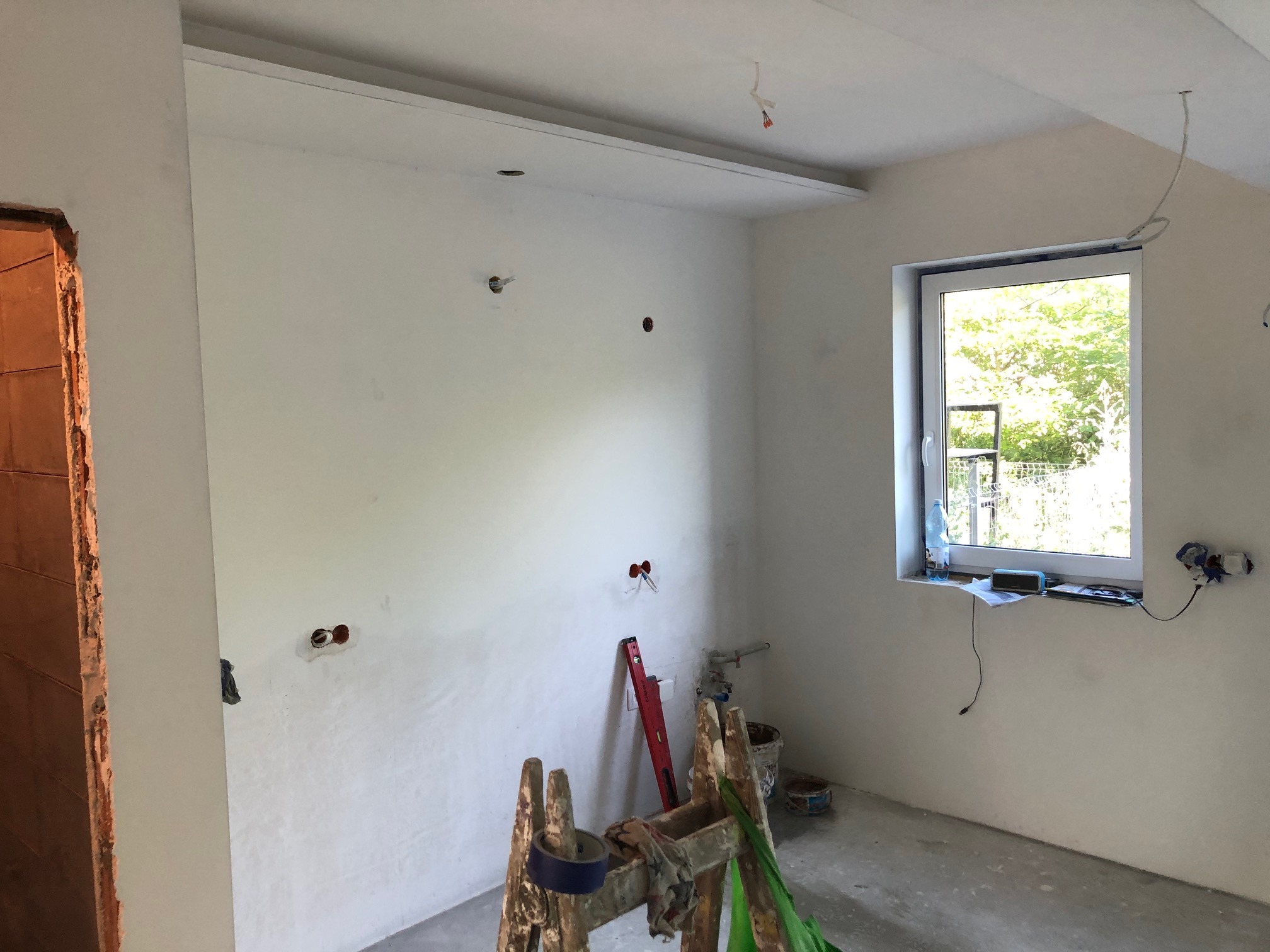 Wnętrze w trakcie remontu: ściany po szpachlowaniu i gruntowaniu, widoczne otwory na instalacje elektryczne, drewniana drabina malarska, okno z widokiem na zieleń.