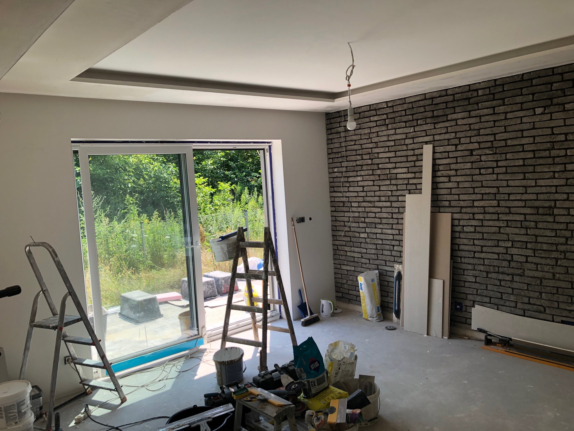 Pokój w trakcie wykańczania: ściana z cegieł, sufit z podwieszaną konstrukcją, okno z widokiem na zieleń, drabiny i worki z materiałami budowlanymi na podłodze.
