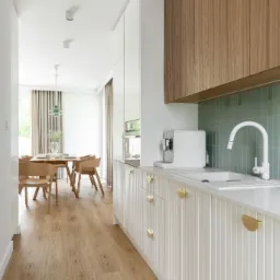 Jasna kuchnia z drewnianymi akcentami, widok na jadalnię z drewnianym stołem i krzesłami, minimalistyczny design, białe meble kuchenne z frezowanymi frontami i złotymi uchwytami, ekspres do kawy...