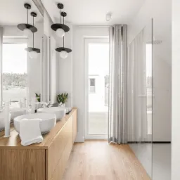 Jasna łazienka z dwoma umywalkami nablatowymi, drewnianą szafką pod umywalkami, kabiną prysznicową ze szklaną ścianą i oknem z widokiem na zieleń za oknem.