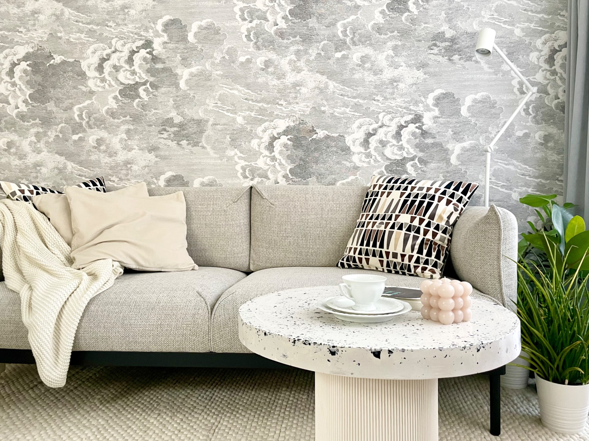 Jasnoszara sofa z poduszkami i pledem na tle tapety w chmury, okrągły stolik kawowy z lastryko z filiżanką, świeczką i rośliną doniczkową, nowoczesna lampa podłogowa.