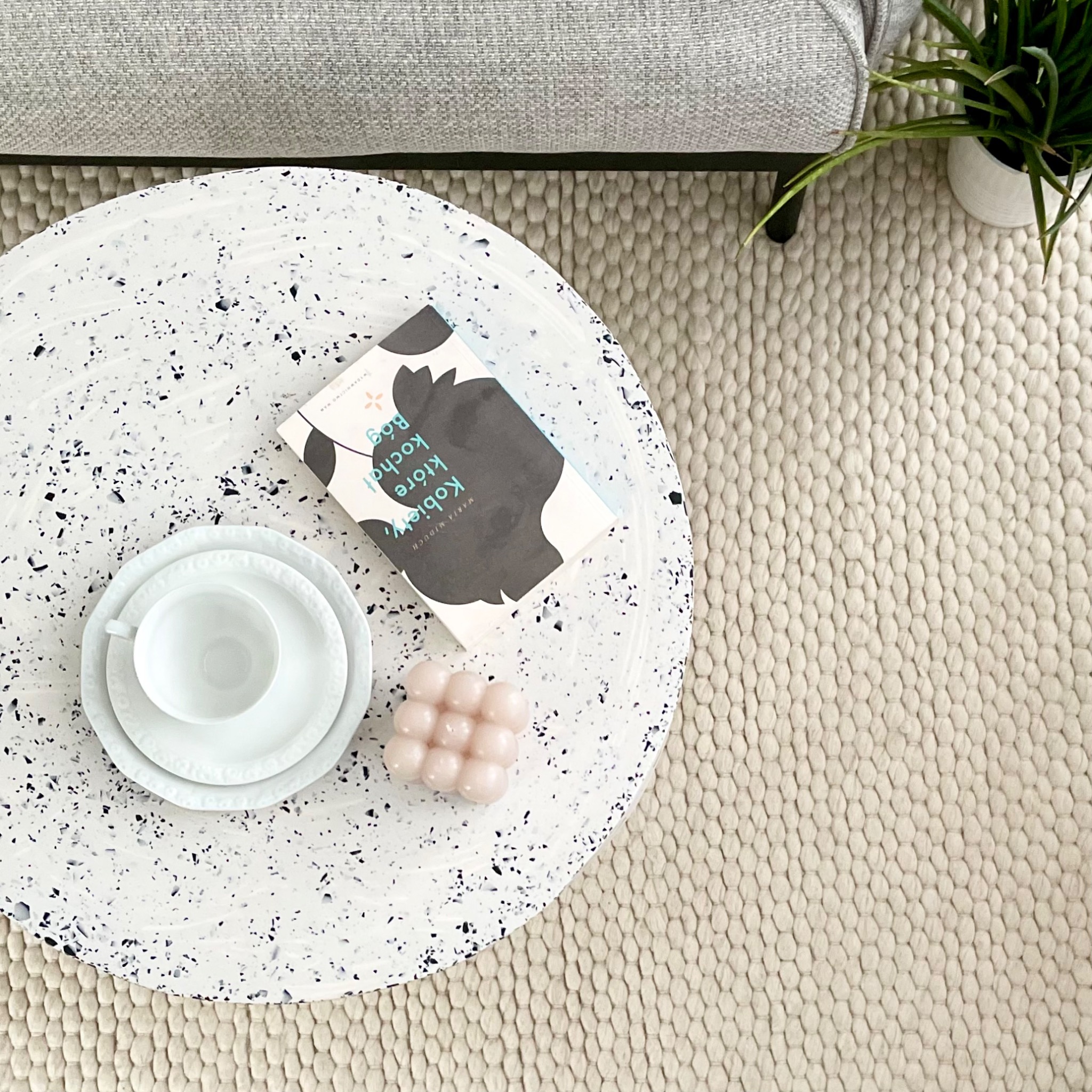 Okrągły stolik kawowy z blatem w stylu lastryko, na którym leżą biała filiżanka ze spodkiem, książka i świeczka bąbelkowa, obok puszysty dywan w odcieniach beżu i szarości oraz fragment sofy...