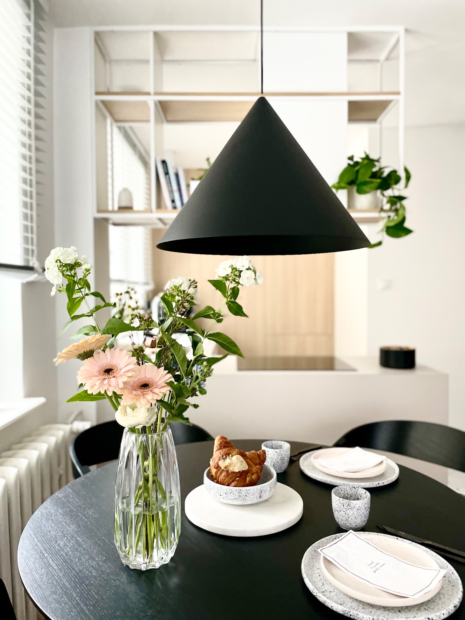 Stół z czarnym blatem, nakryty porcelanową zastawą i ozdobiony bukietem kwiatów w szklanym wazonie, nad stołem wisi czarna lampa w kształcie stożka, w tle białe półki z książkami i roślinami.