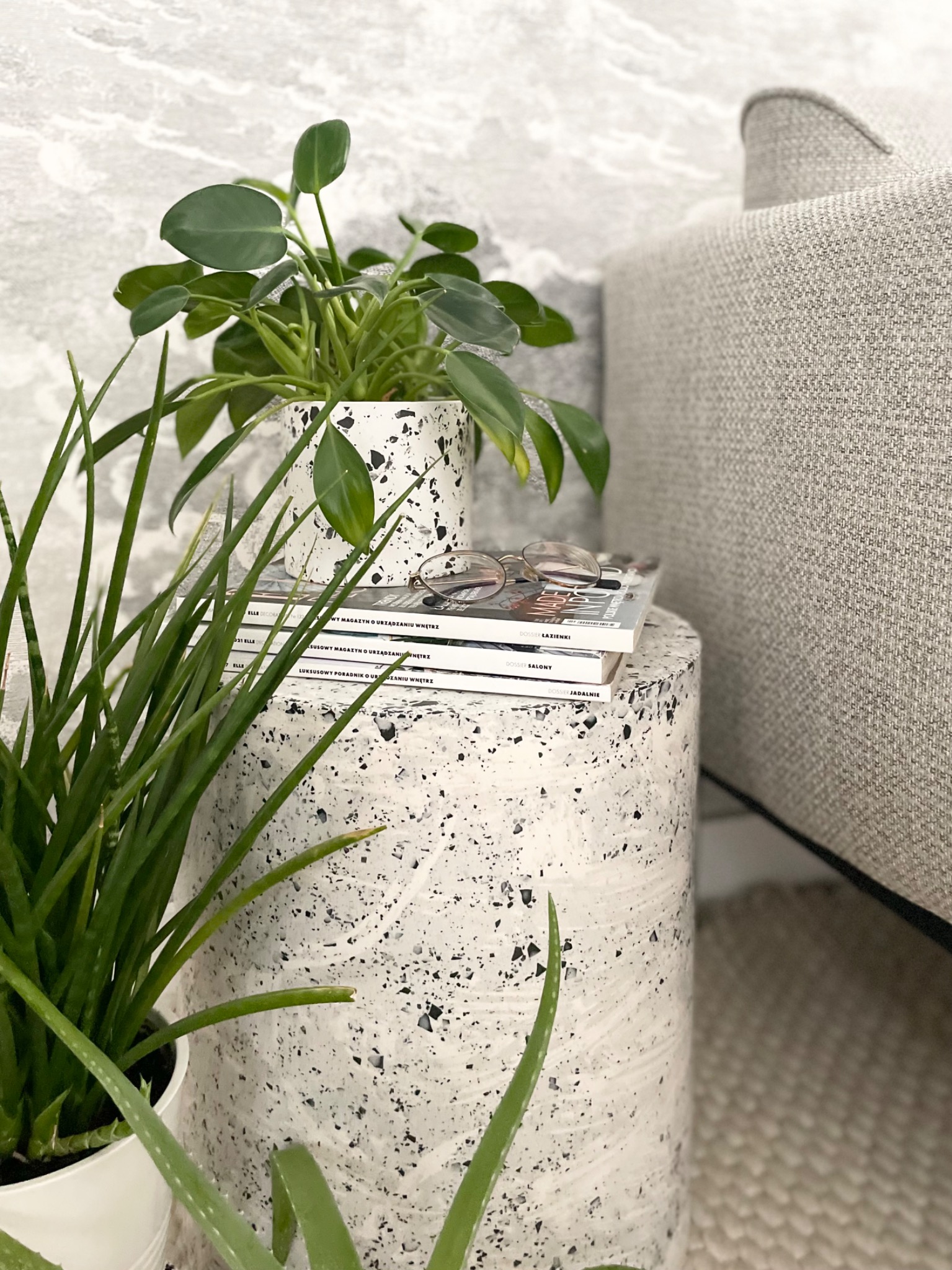 Stolik pomocniczy z terazzo, na nim ułożone magazyny wnętrzarskie, okulary i doniczka z rośliną, obok sofa w jasnej tapicerce i doniczka z sansewierią, całość w stonowanej, jasnej kolorystyce.