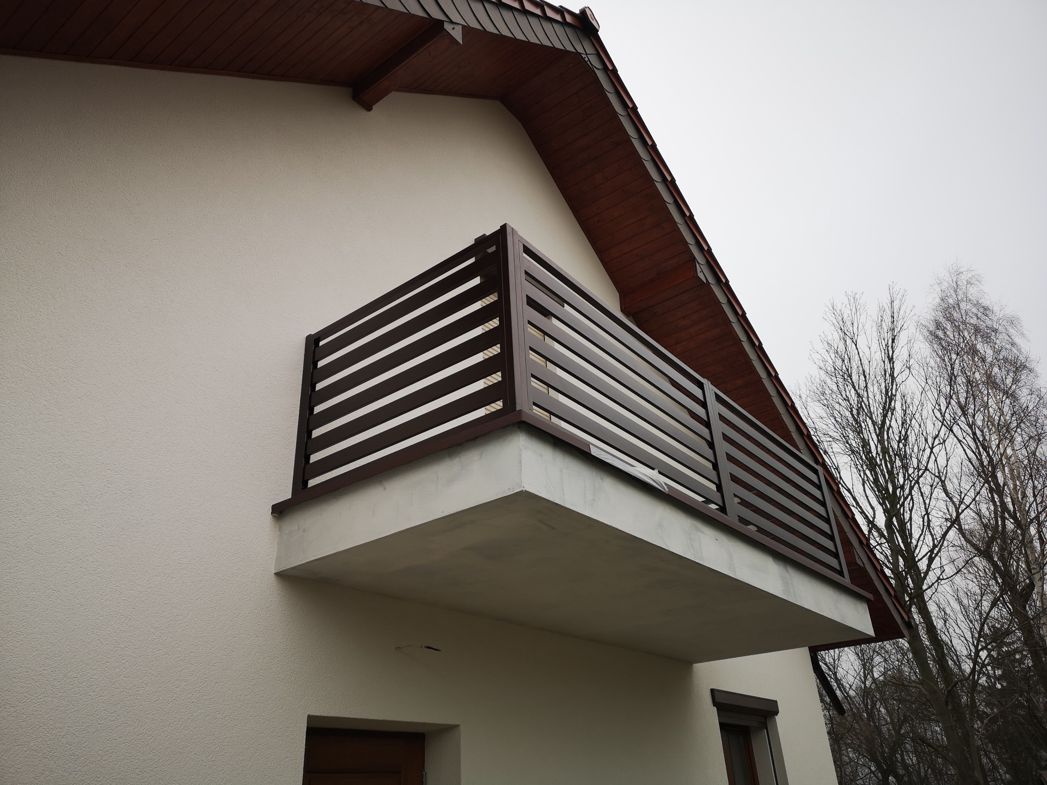 Nowoczesna balustrada balkonowa z poziomych, brązowych profili na tle białej elewacji domu z brązowym dachem i drzewami bez liści.