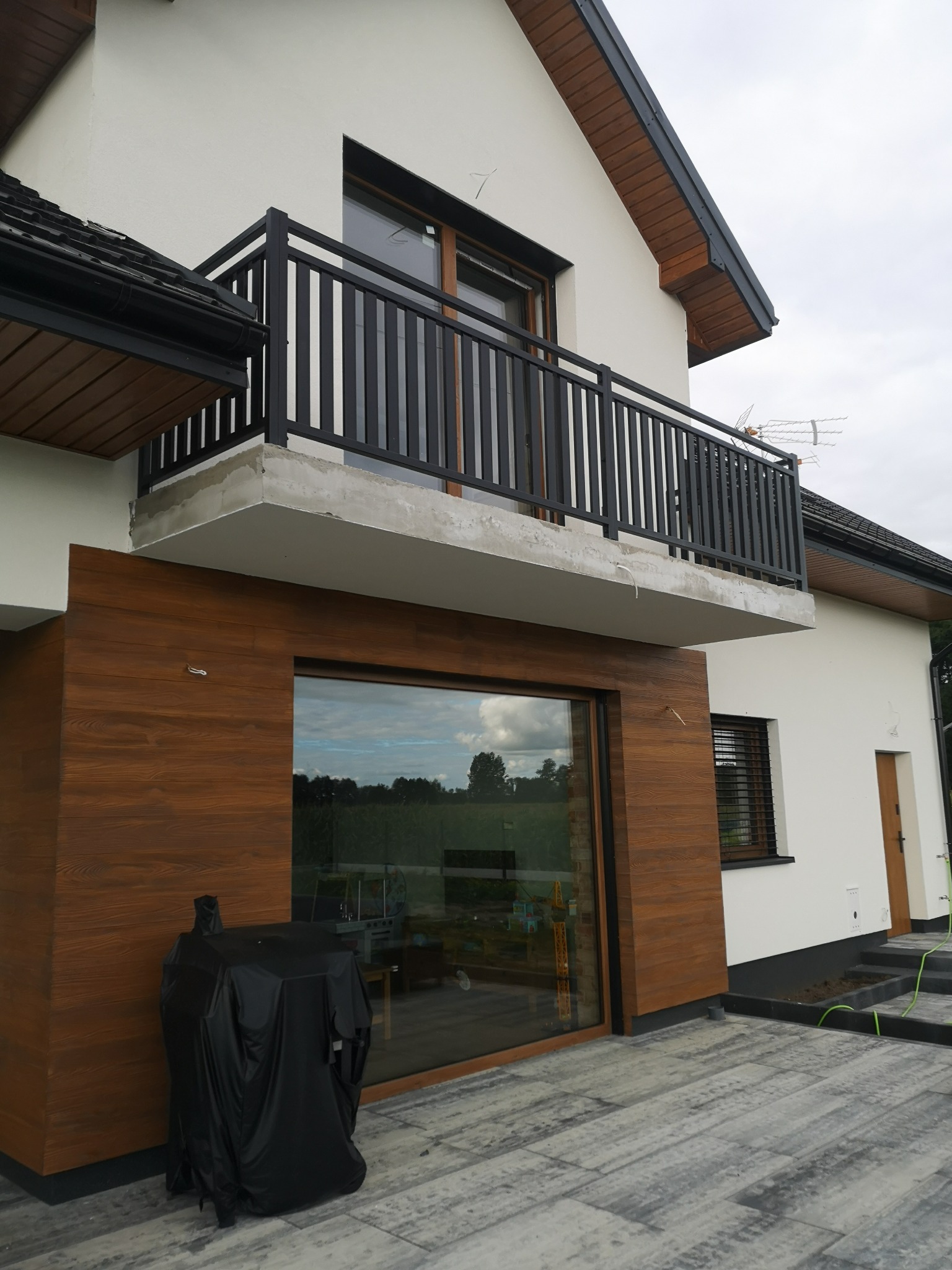 Nowoczesny dom z balkonem z czarną, pionową balustradą, drewnianą elewacją i dużym oknem, na pierwszym planie grill pod pokrowcem.