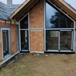 Wind-Best S.C. Dawid Grabowski Sylwia Grabowska - Surowy stan domu w trakcie budowy z zamontowanymi oknami PCV o czarnych ramach, widoczne cegły, drewniane elementy konstrukcji dachu i błotniste podłoże przed budynkiem.