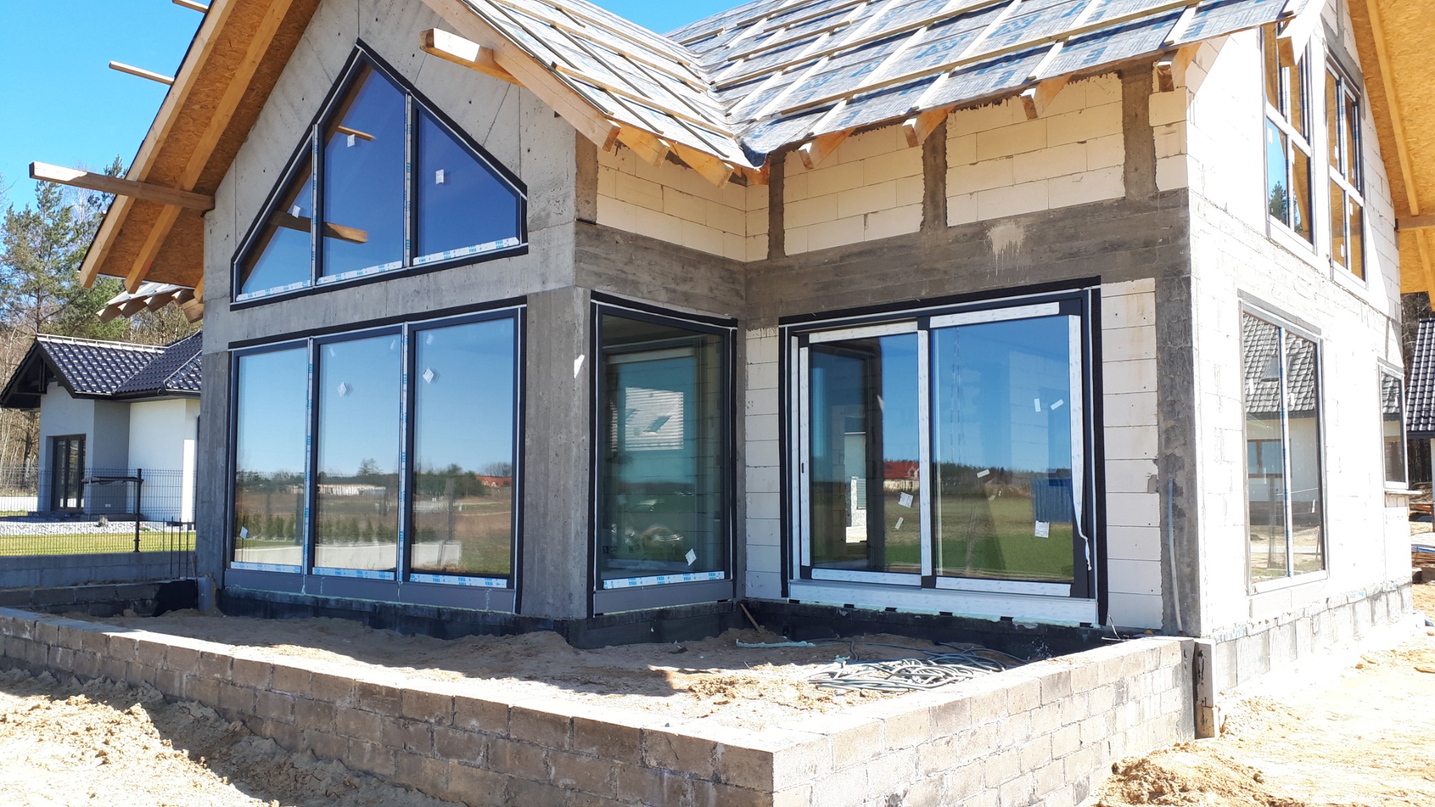 Surowy stan domu w trakcie budowy z zamontowanymi oknami PCV z czarnymi ramami i dużymi przeszkleniami, widoczne niezabezpieczone kable na fundamencie.