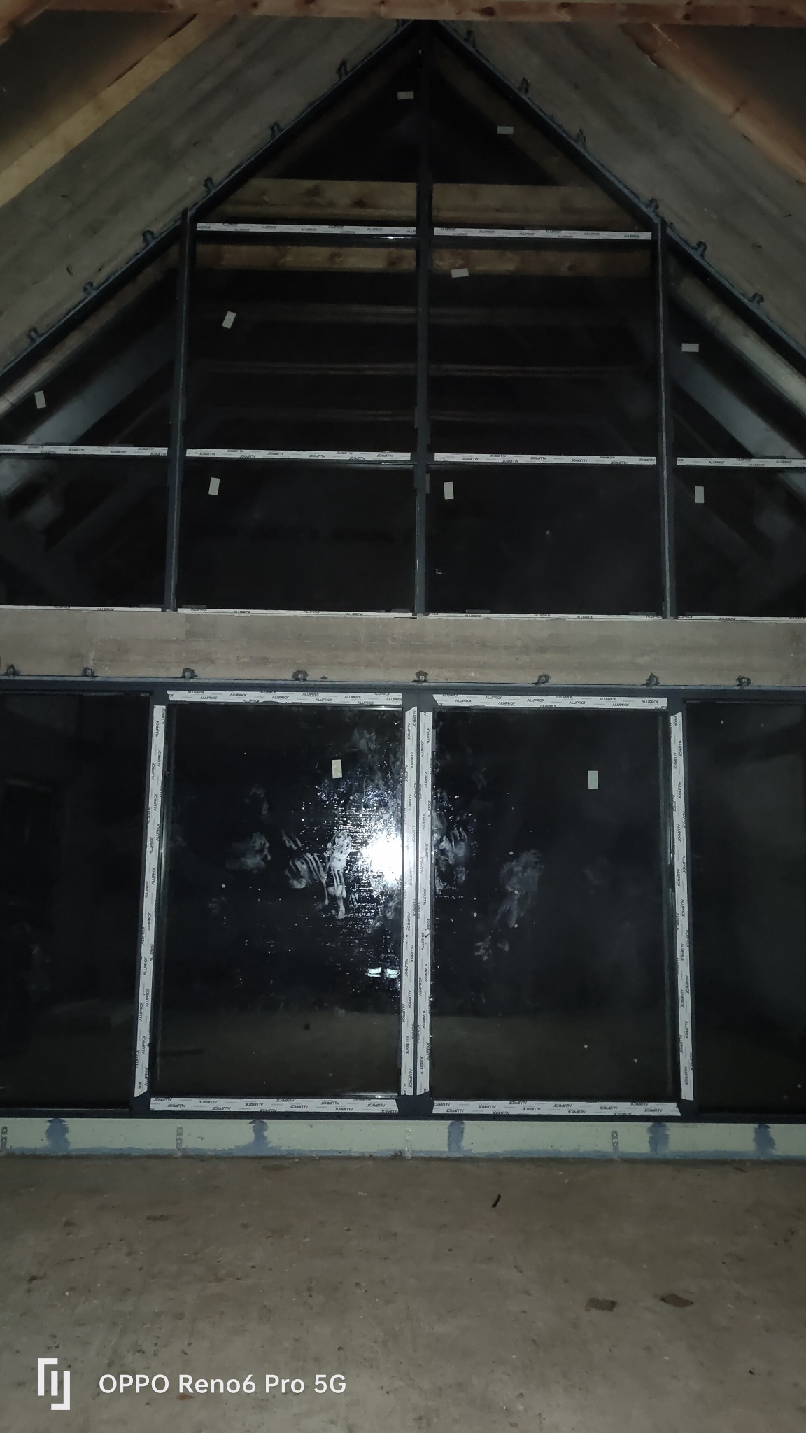 Montaż okien w surowym stanie poddasza, widoczne ramy okienne z szybami, betonowe nadproże i drewniana konstrukcja dachu.