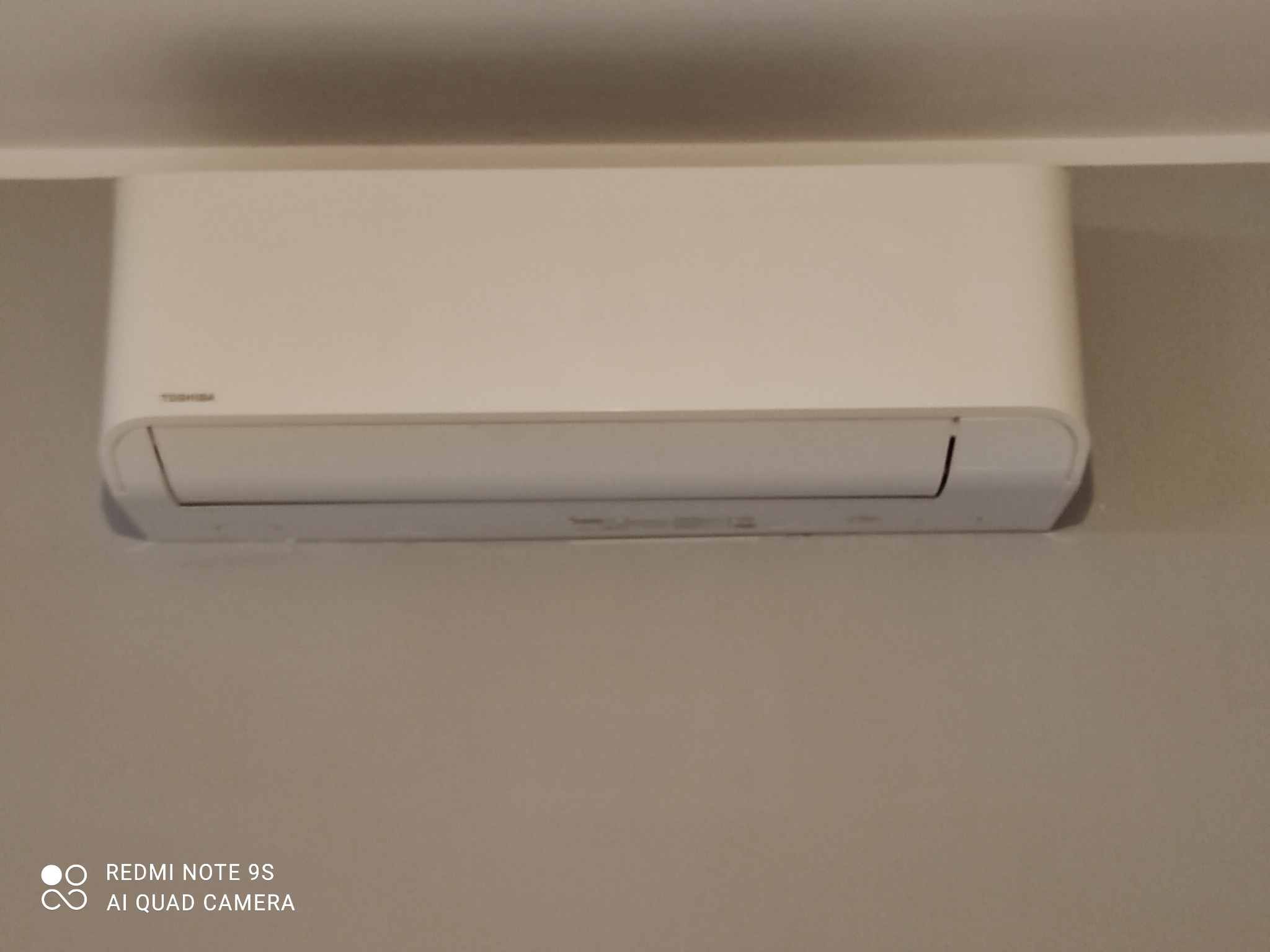 Biały klimatyzator Toshiba zamontowany na szarej ścianie w pomieszczeniu.