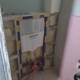 Stelaż podtynkowy Grohe zamontowany w konstrukcji z profili i izolacji z wełny mineralnej, zabezpieczony żółtą taśmą, widoczne elementy instalacji hydraulicznej.