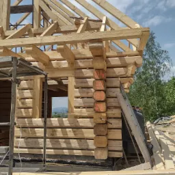 Surowa konstrukcja domu z bali w trakcie budowy, widoczne rusztowania i elementy więźby dachowej, w tle zielone drzewa i błękitne niebo.