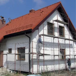 Dom jednorodzinny w trakcie ocieplania elewacji, widoczne rusztowania i pracownicy, fragment otynkowanej ściany przy gruncie, dach pokryty czerwoną dachówką.