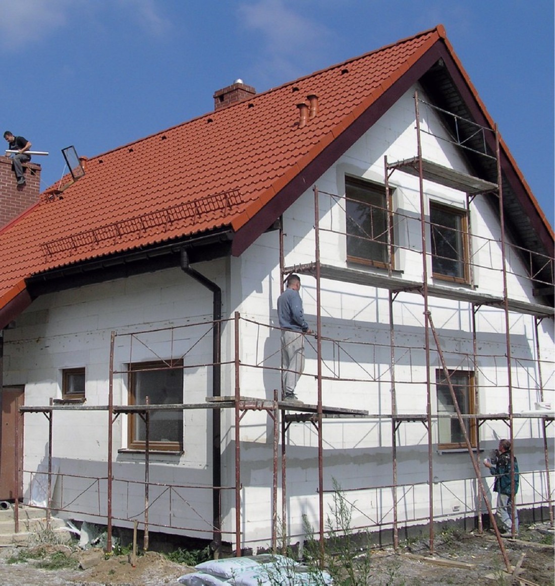 Dom jednorodzinny w trakcie ocieplania elewacji, widoczne rusztowania i pracownicy, fragment otynkowanej ściany przy gruncie, dach pokryty czerwoną dachówką.