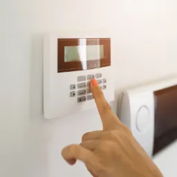 Dłoń z pomalowanym na koralowo paznokciem naciska przycisk '2' na białym panelu sterowania alarmem domowym zamontowanym na białej ścianie. W tle widoczny biały termostat i okulary.