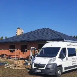 Nowo pokryty, ciemny dach z blachodachówki na surowym budynku w trakcie budowy, z widocznymi rusztowaniami i stertą materiałów dekarskich na trawie, na tle jasnego, błękitnego nieba i firmowego...