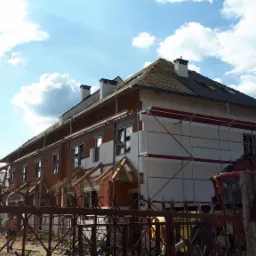 Budynek w trakcie ocieplania, widoczne rusztowania, częściowo położona dachówka, przygotowane drewniane elementy konstrukcyjne nad oknami, styropianowe płyty izolacyjne na elewacji.