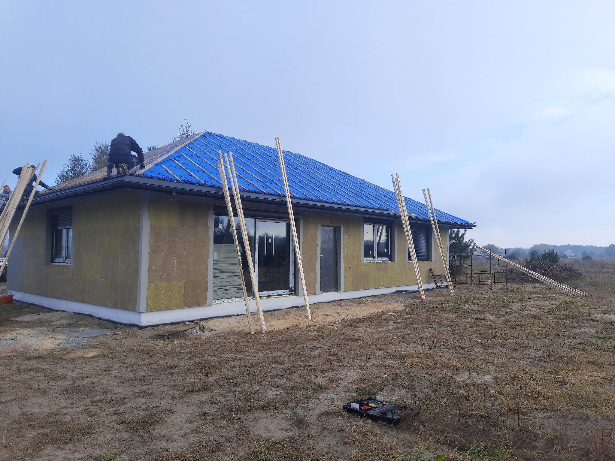 Budowa domu jednorodzinnego: Ekipa montuje niebieską membranę dachową na nowym dachu. Ocieplenie ścian zewnętrznych w trakcie, widoczne narzędzia i rusztowania.