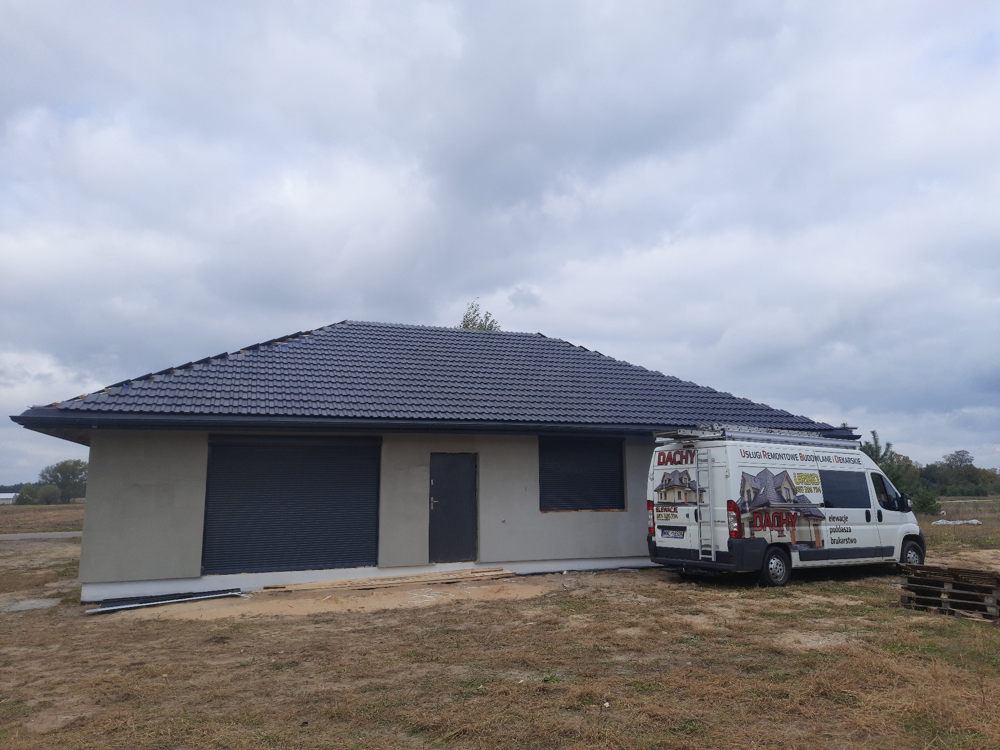 Nowoczesny dom jednorodzinny z ciemnoszarą dachówką ceramiczną, szarymi roletami zewnętrznymi i jasnoszarą elewacją, obok zaparkowany biały samochód dostawczy z reklamą firmy dekarskiej.