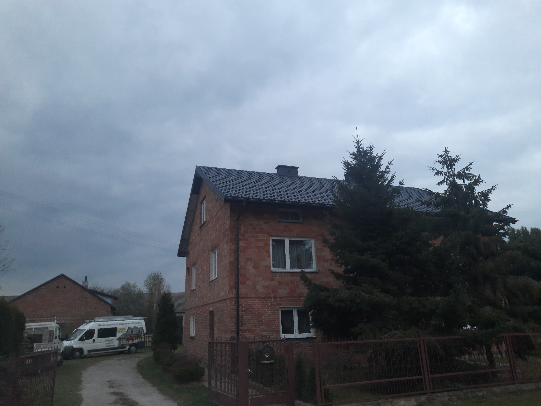 Dwupiętrowy dom z czerwonej cegły z ciemnoszarą dachówką, widoczny zza ogrodzenia, z firmowym vanem dekarza zaparkowanym na podjeździe.