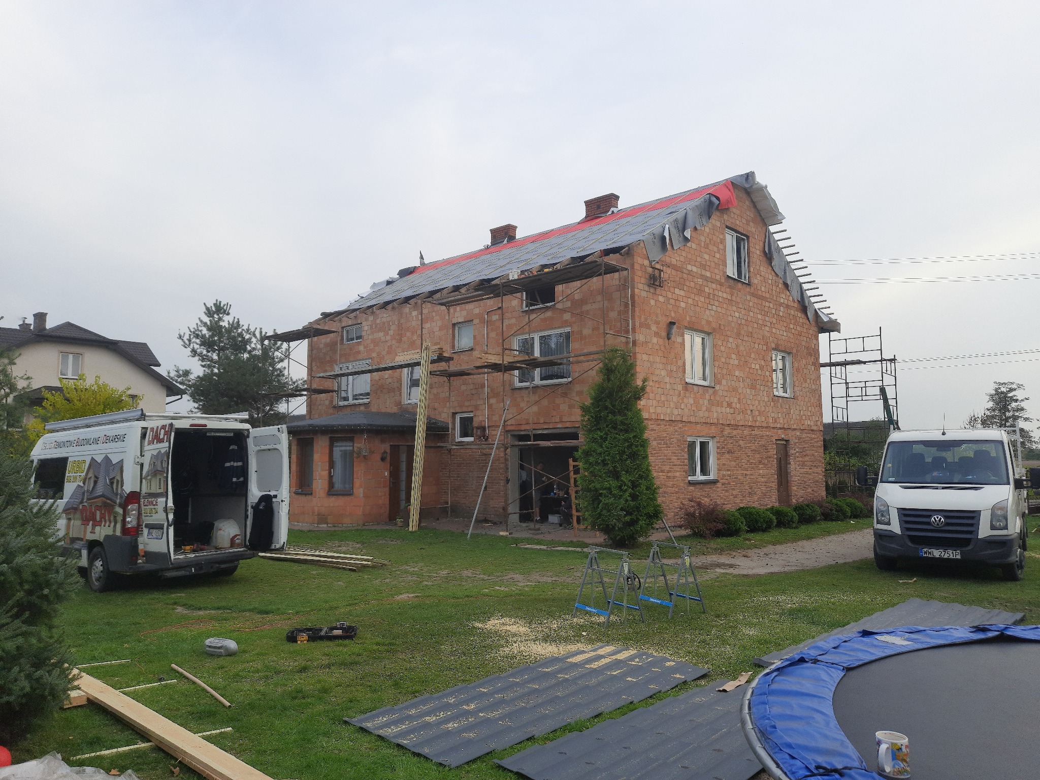 Dwupiętrowy dom z cegły w trakcie wymiany dachu, zabezpieczony folią, z rusztowaniami i dwoma samochodami dostawczymi firmy dekarskiej na posesji.
