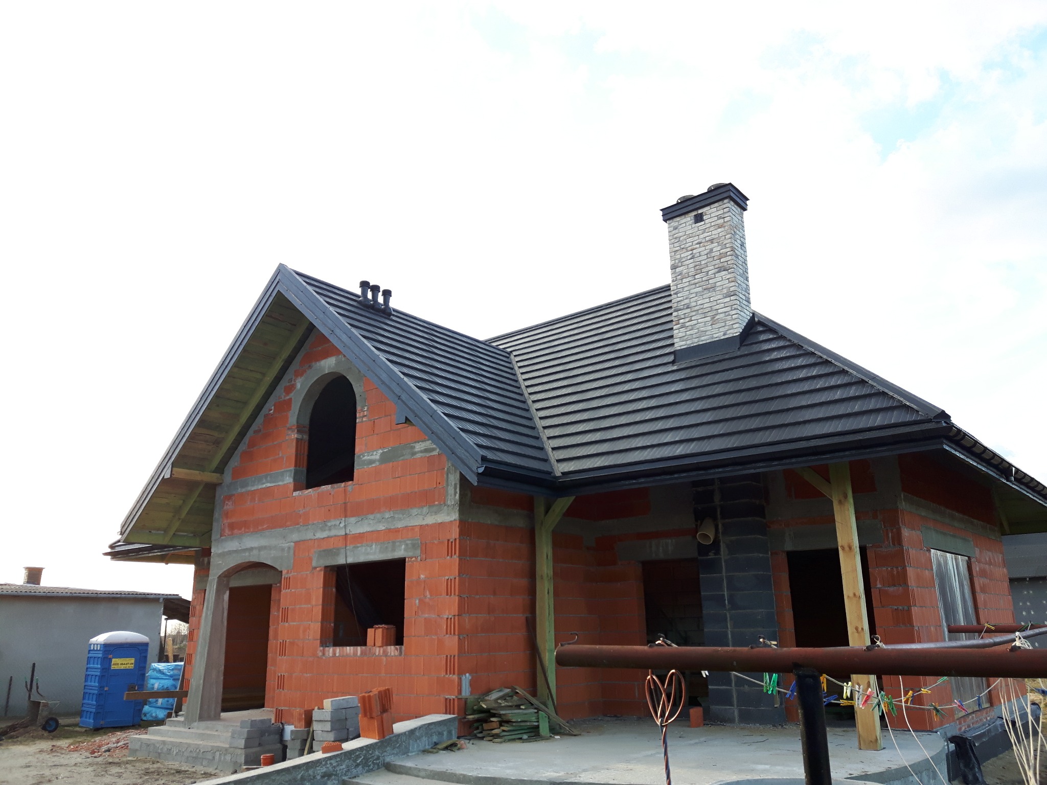 Dom w trakcie budowy z widocznym dachem pokrytym ciemną dachówką, murowanym kominem i ścianami z czerwonej cegły. Widoczne także elementy konstrukcyjne i tymczasowe zabezpieczenia.