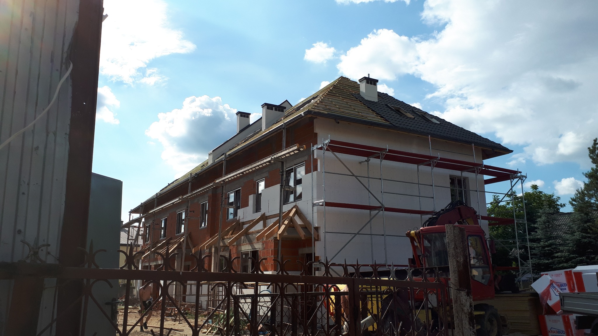 Budynek w trakcie ocieplania, widoczne rusztowania, częściowo położona dachówka, przygotowane drewniane elementy konstrukcyjne nad oknami, styropianowe płyty izolacyjne na elewacji.