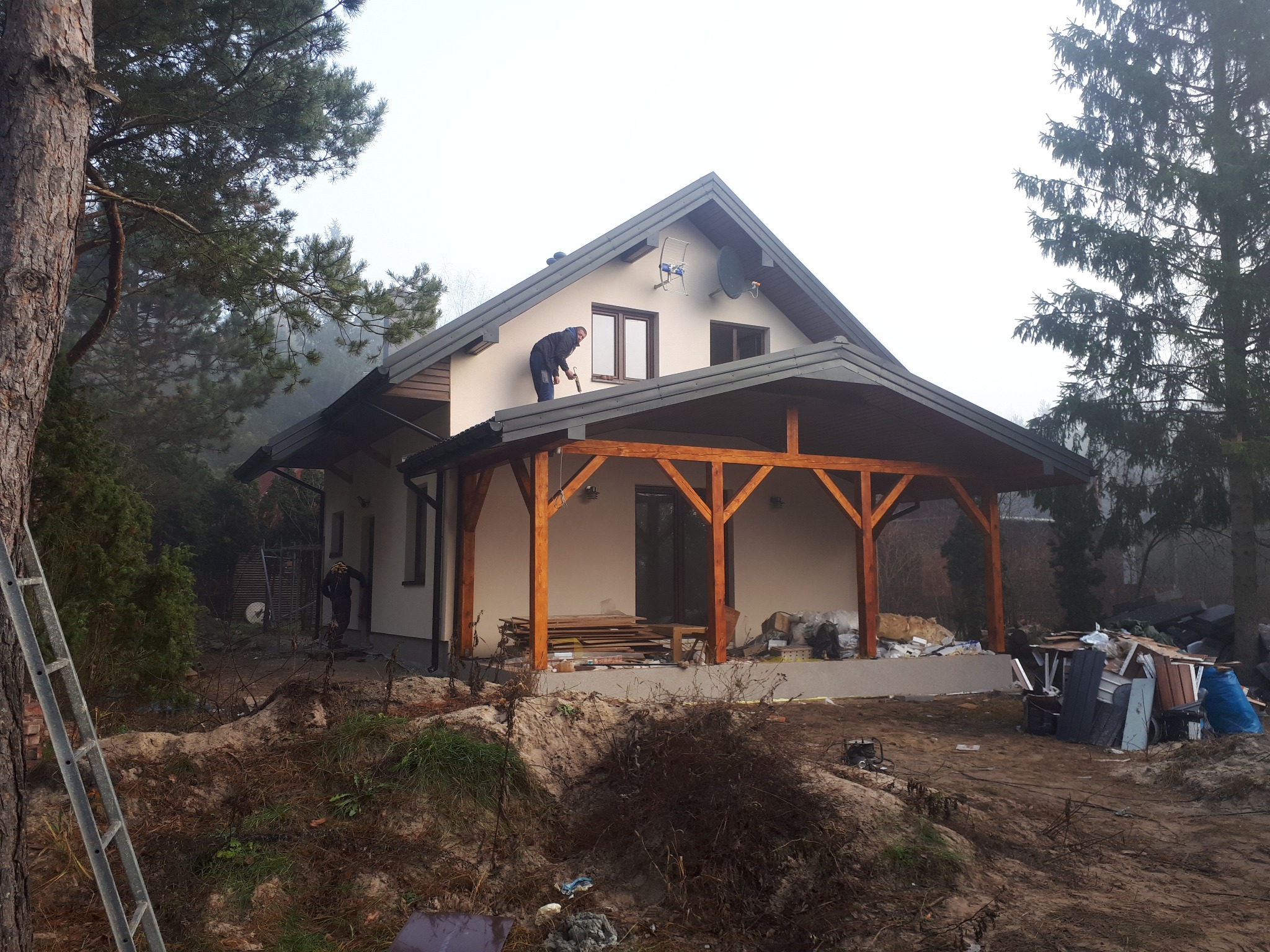 Dom jednorodzinny z szarą dachówką, na dachu pracownik z narzędziem, widoczna drewniana konstrukcja zadaszenia tarasu, teren wokół domu w trakcie prac wykończeniowych.
