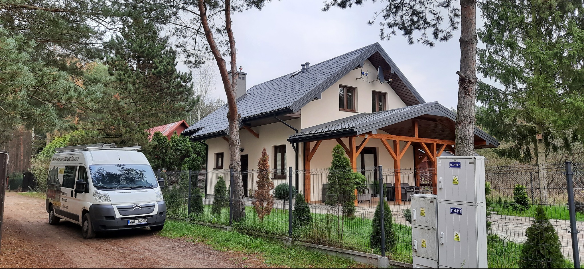 Dom jednorodzinny z szarą dachówką karpiówką i obróbkami blacharskimi, widoczny zza ogrodzenia i drzew, z zaparkowanym białym busem firmy dekarskiej.