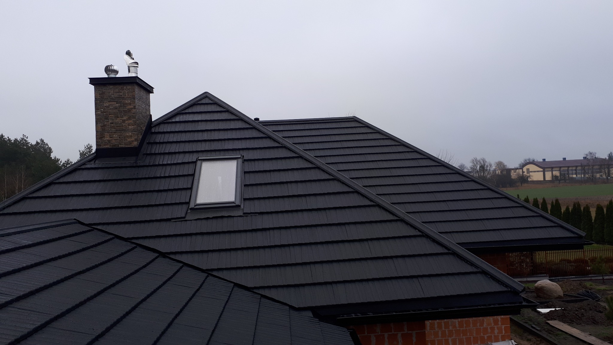 Czarny dach z dachówki modułowej z oknem połaciowym i kominem obłożonym kamieniem dekoracyjnym, w tle zalesiony teren i budynek gospodarczy.