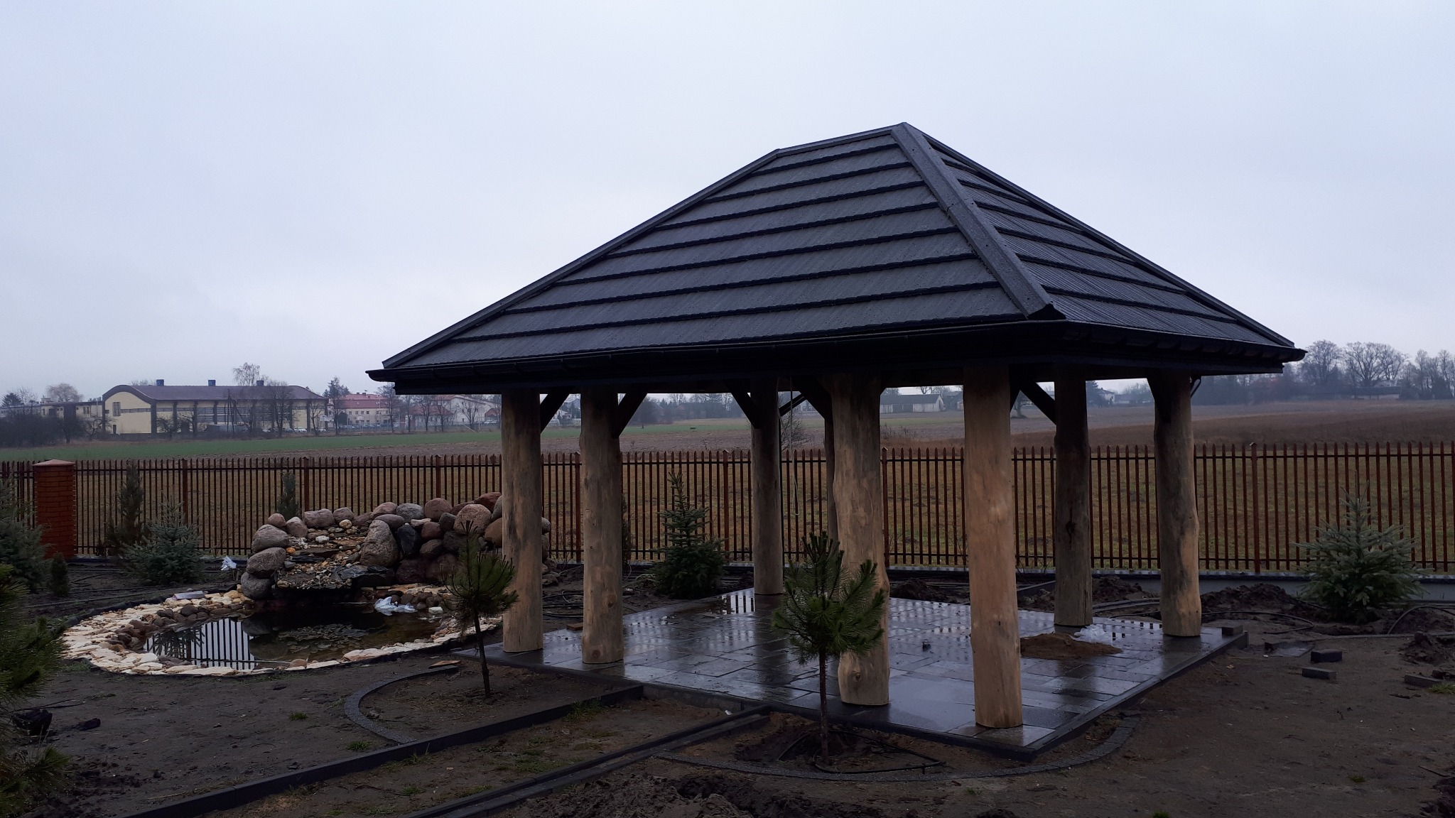Altana z ciemnym, metalowym dachem z widoczną strukturą paneli, wsparta na surowych, drewnianych słupach, obok oczko wodne z kaskadą, w tle brązowe ogrodzenie i budynki.
