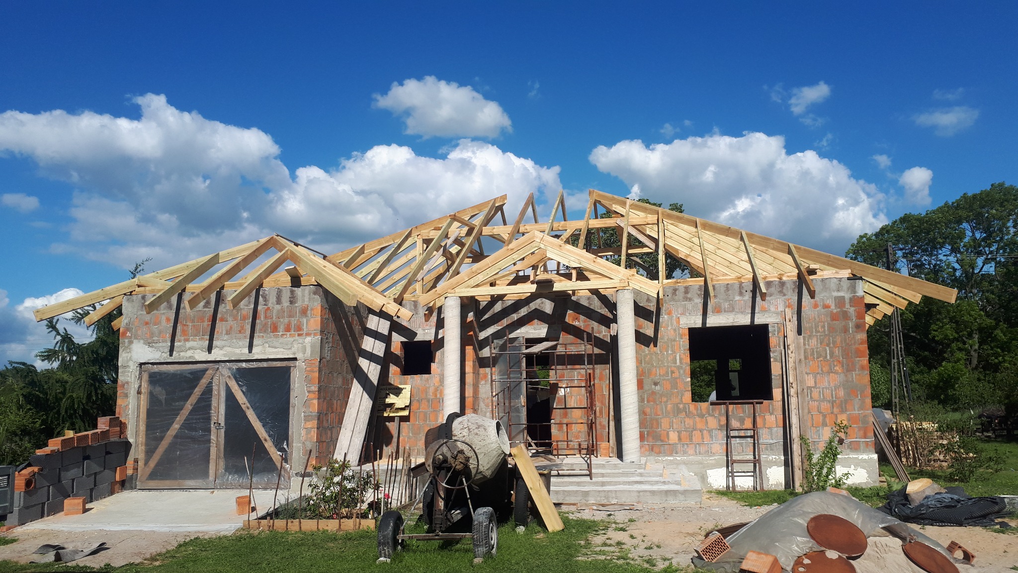 Budynek w trakcie budowy z widoczną konstrukcją drewnianą dachu, betoniarką i elementami rusztowania na tle błękitnego nieba z chmurami.